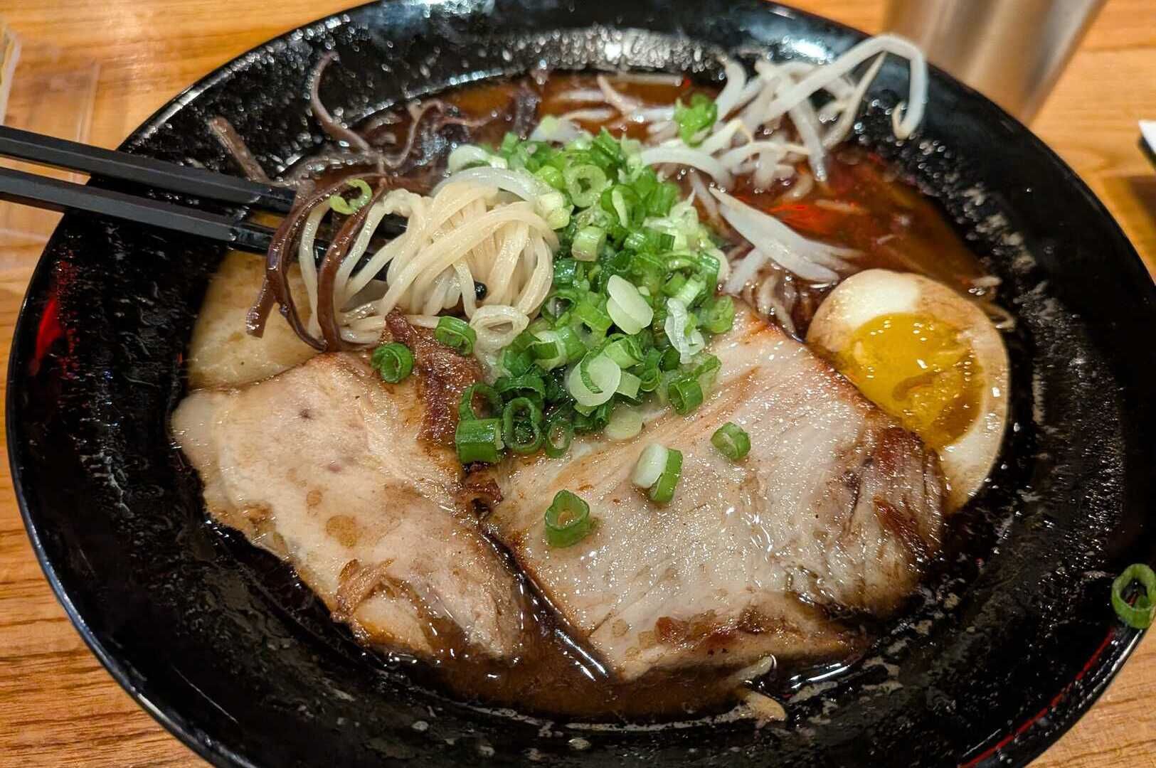 A bowl of Tonkotsu Black Ramen topped with meat and fresh vegetables, showcasing rich broth and vibrant colors.