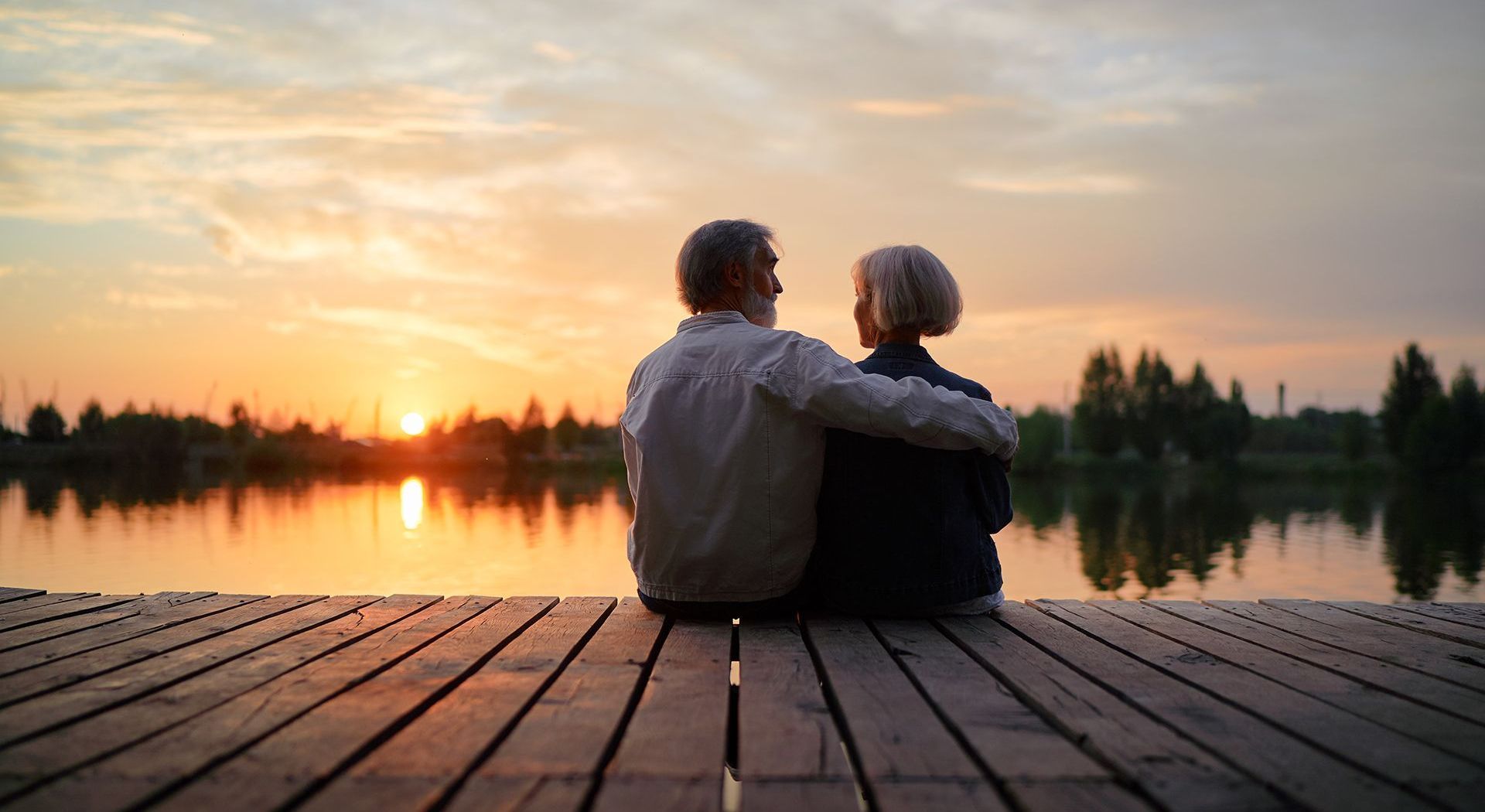 An elderly couple is sitting on a dock looking at the sunset over a lake.