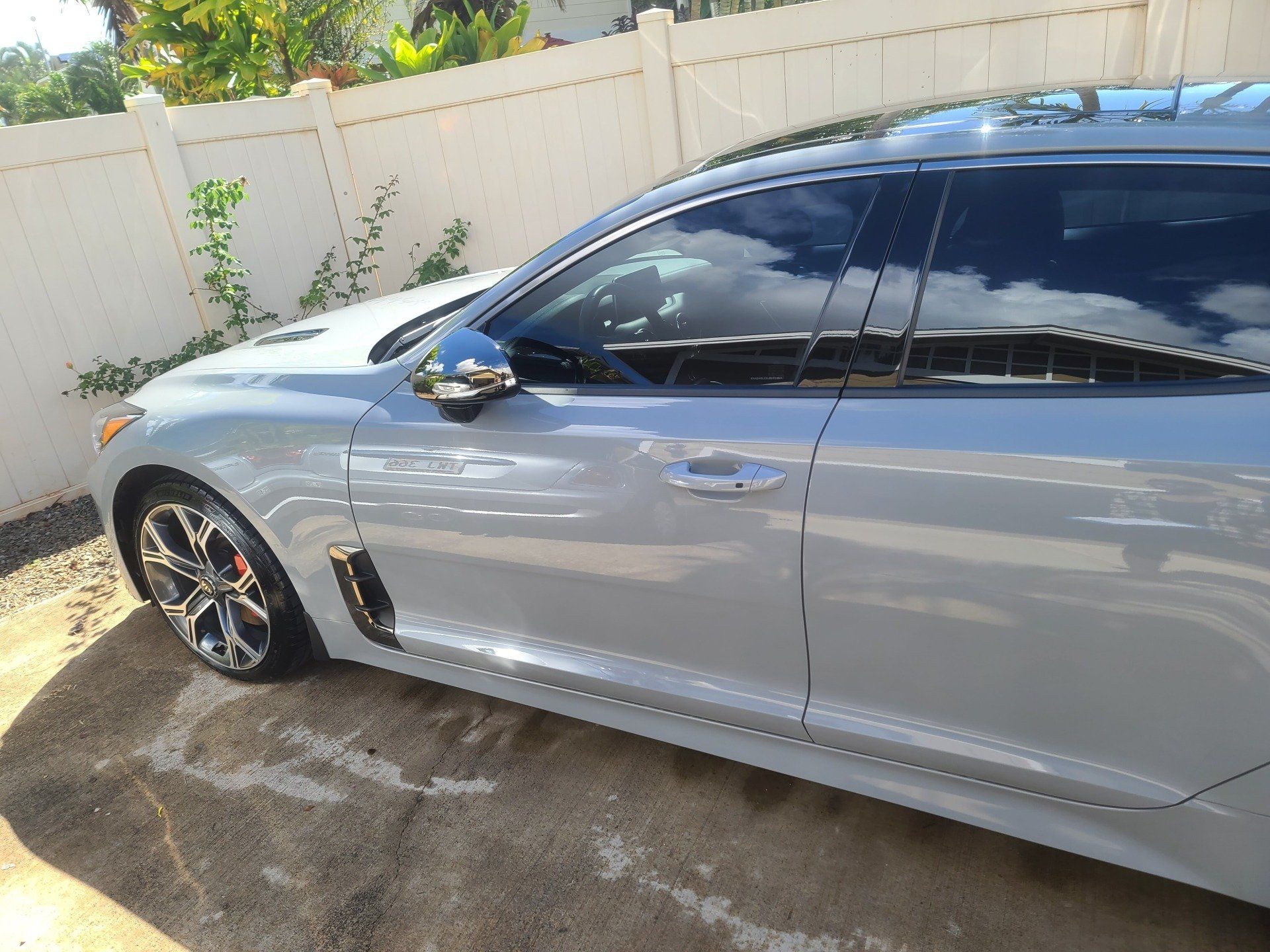 Silver Kia Stinger parked on a paved surface near a white fence, reflecting light.