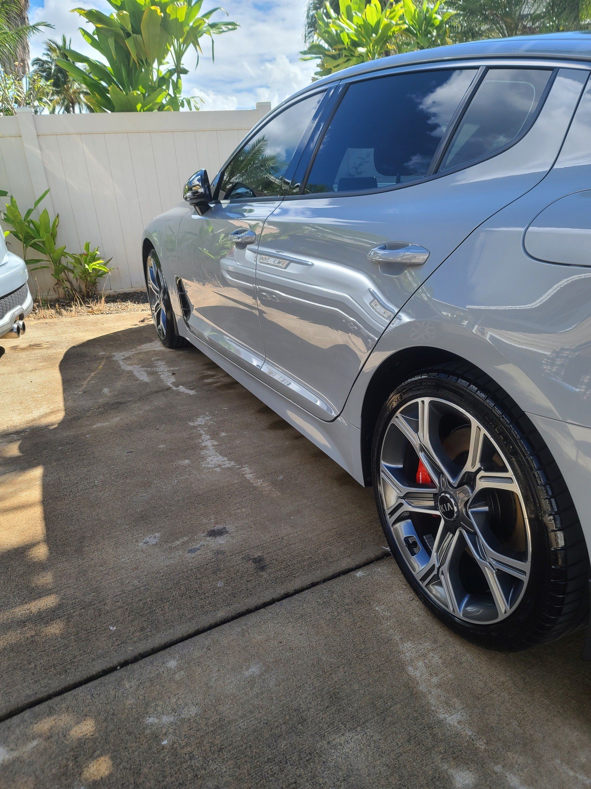 White Kia Stinger parked on a concrete driveway, near a white wall, with a partially visible car.