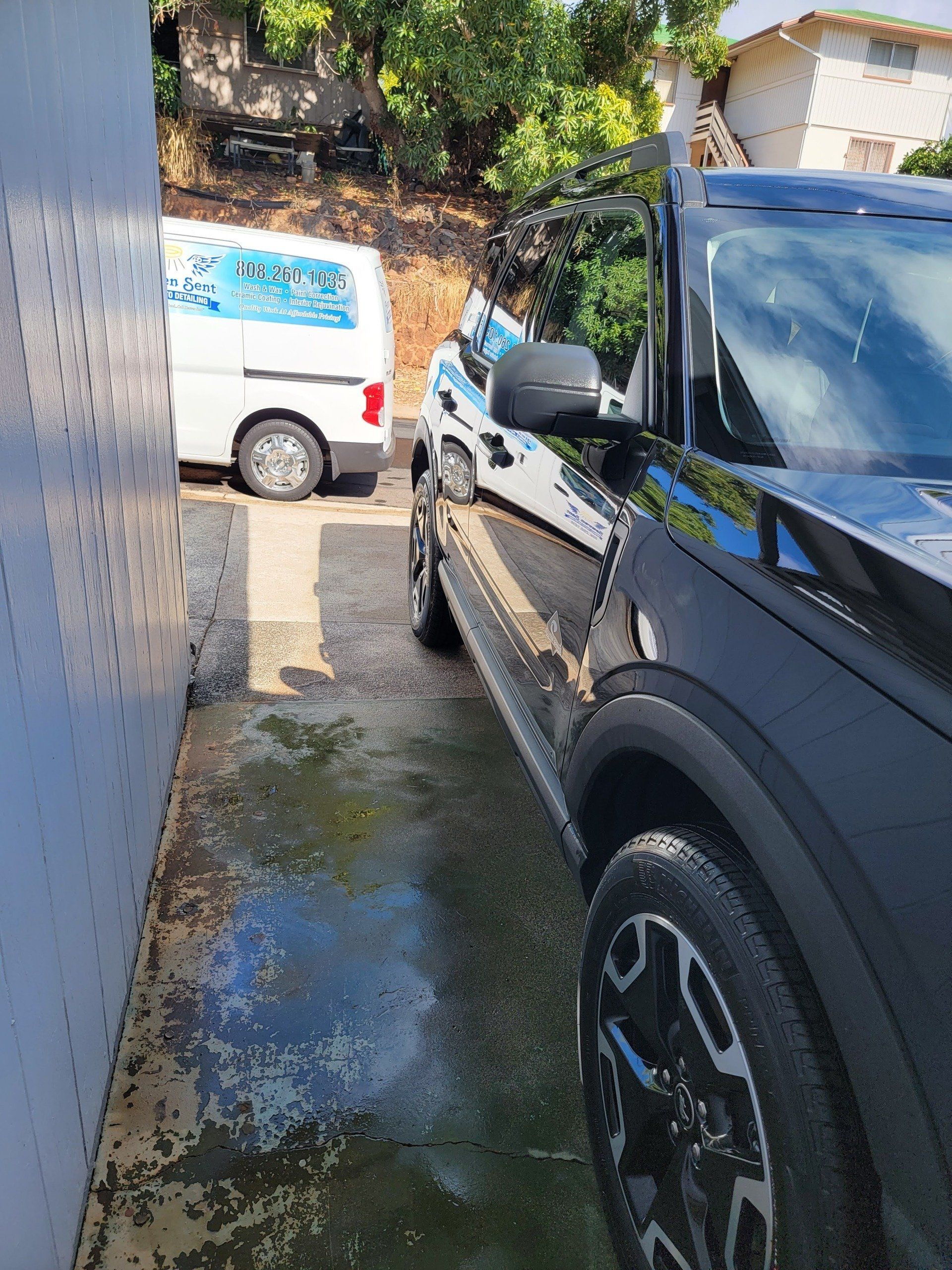 Black SUV parked next to a white van in a narrow alley. Wet ground. Sunny day.
