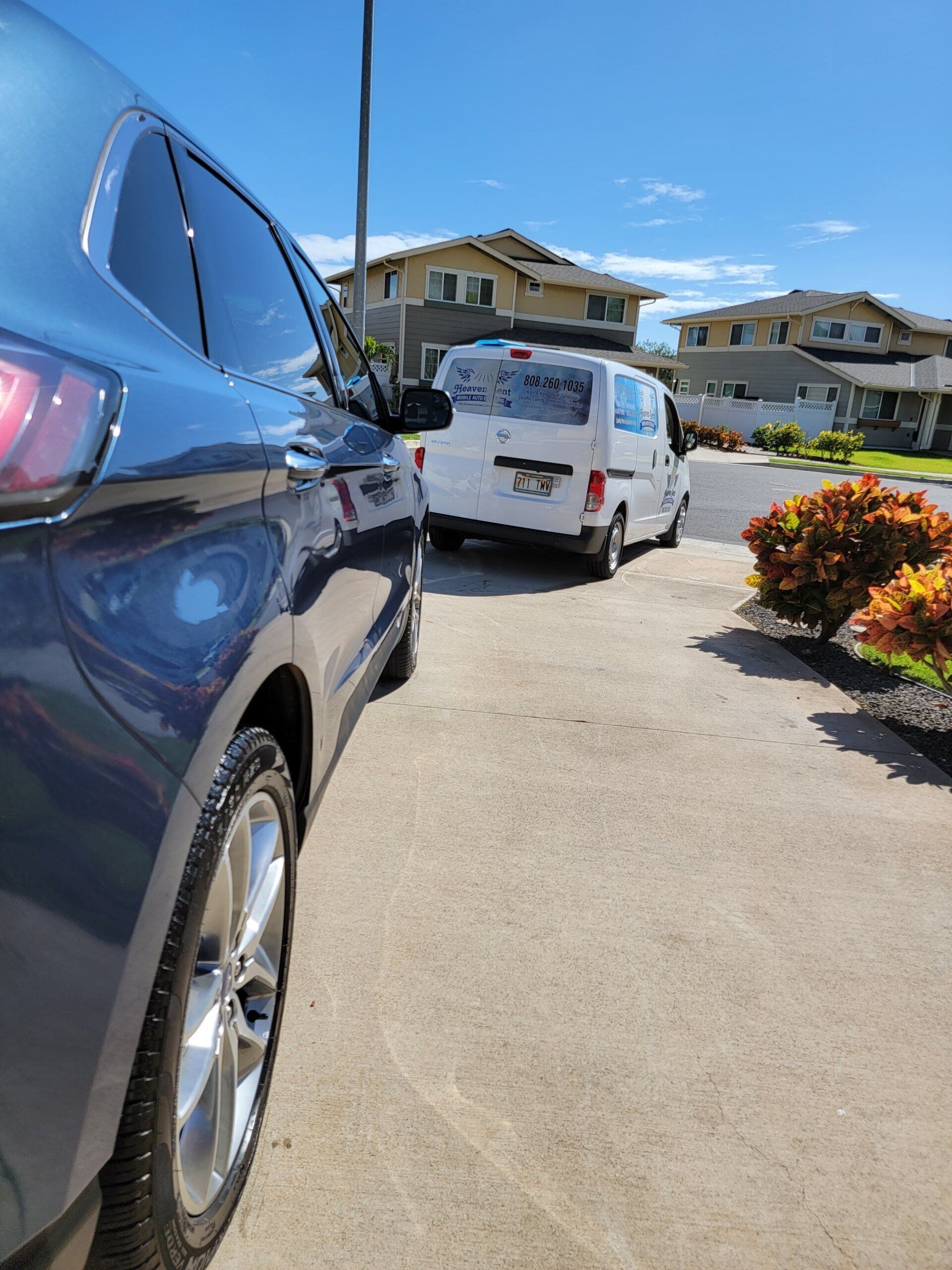 Blue car parked on driveway, white van in background, sunny day.