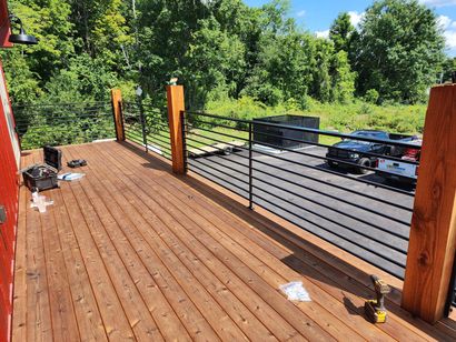 Elevated wooden deck with cable railing, orange wood posts, overlooking a forested area and parking lot below - NRK Welding LLC