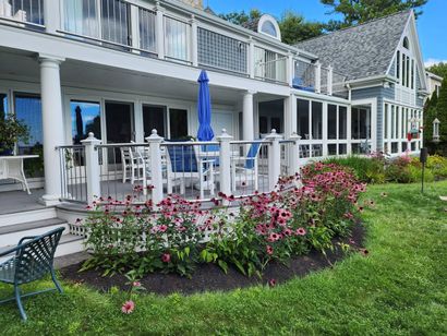 Coastal home with wraparound porch, white columns, blue umbrellas, and vibrant pink flower bed in the front yard - NRK Welding LLC