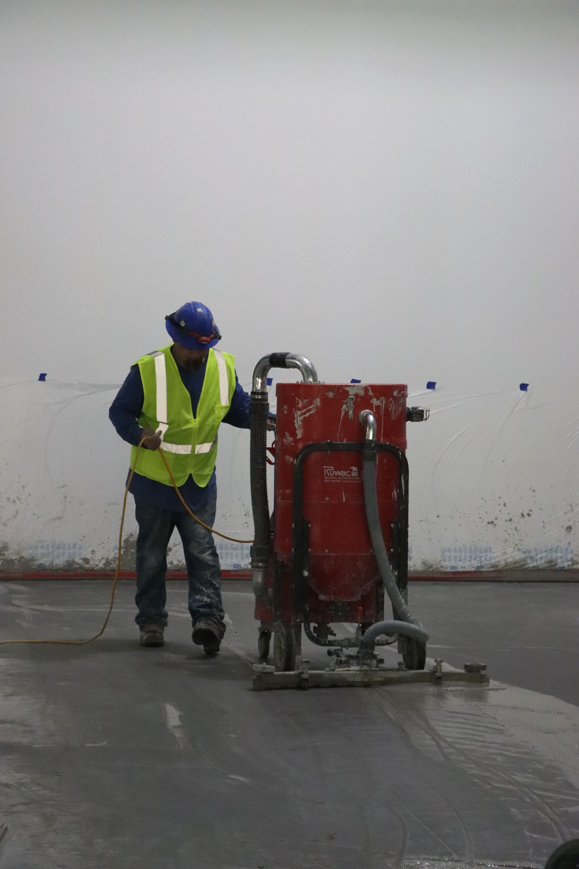 A construction worker is using a vacuum cleaner on a concrete floor.