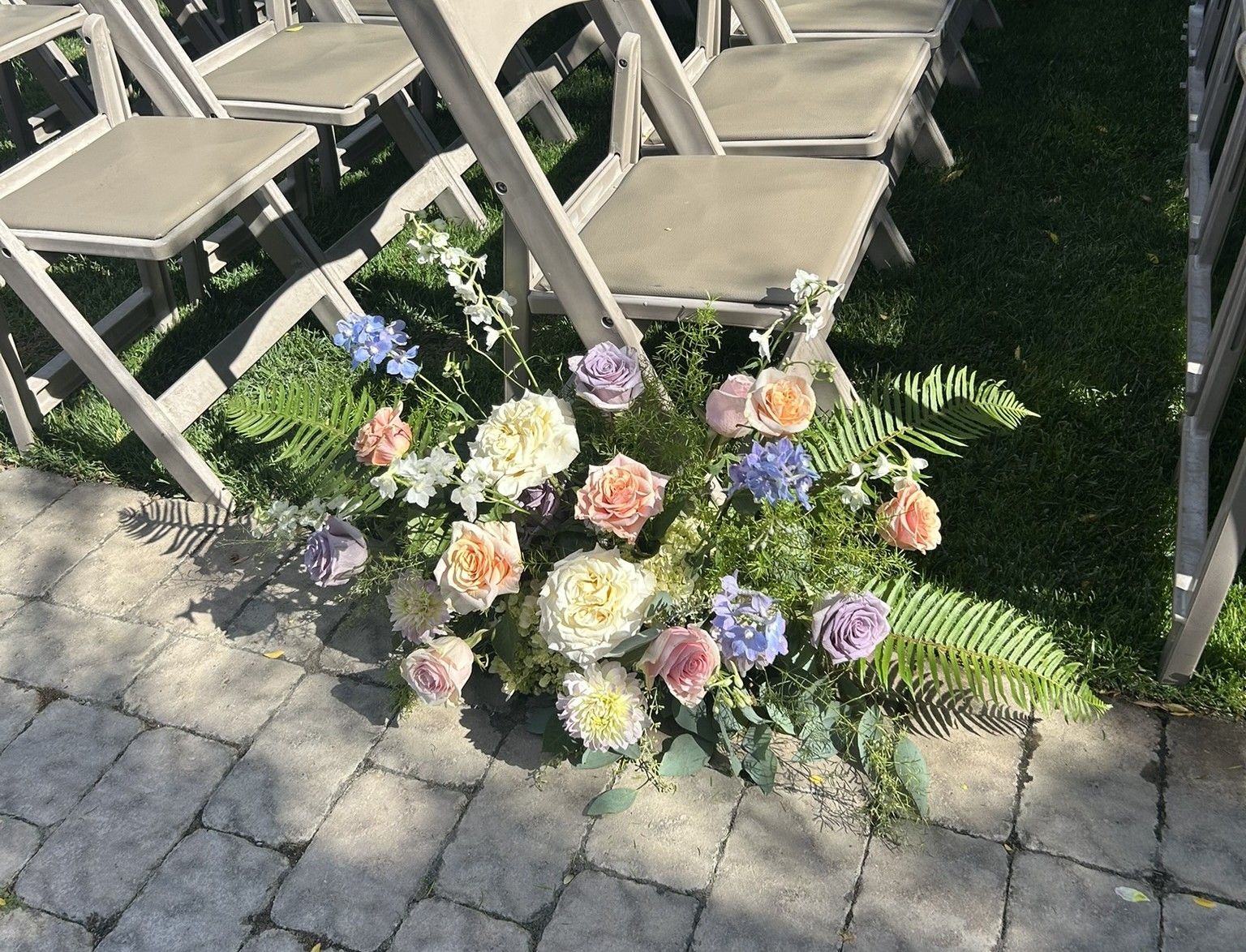 Floral arrangement at the base of chairs in a wedding setting: pastel flowers, ferns, gray chairs, brick and grass.