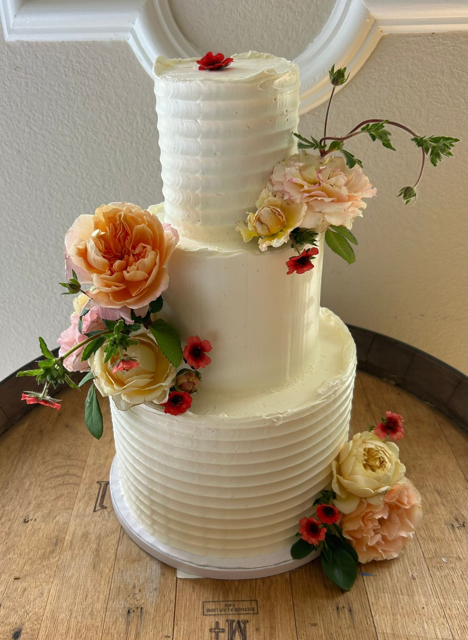 A white wedding cake with flowers on it is sitting on top of a wooden barrel.