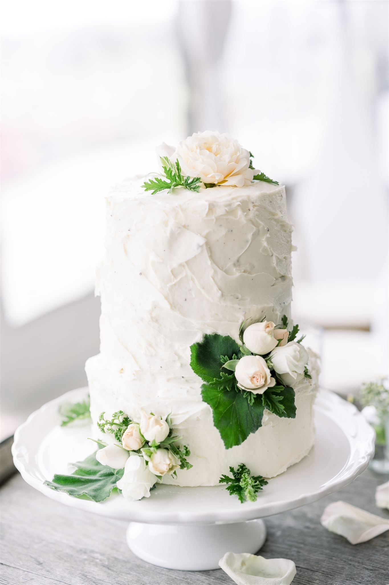 A white wedding cake with white flowers and green leaves on a white cake stand.