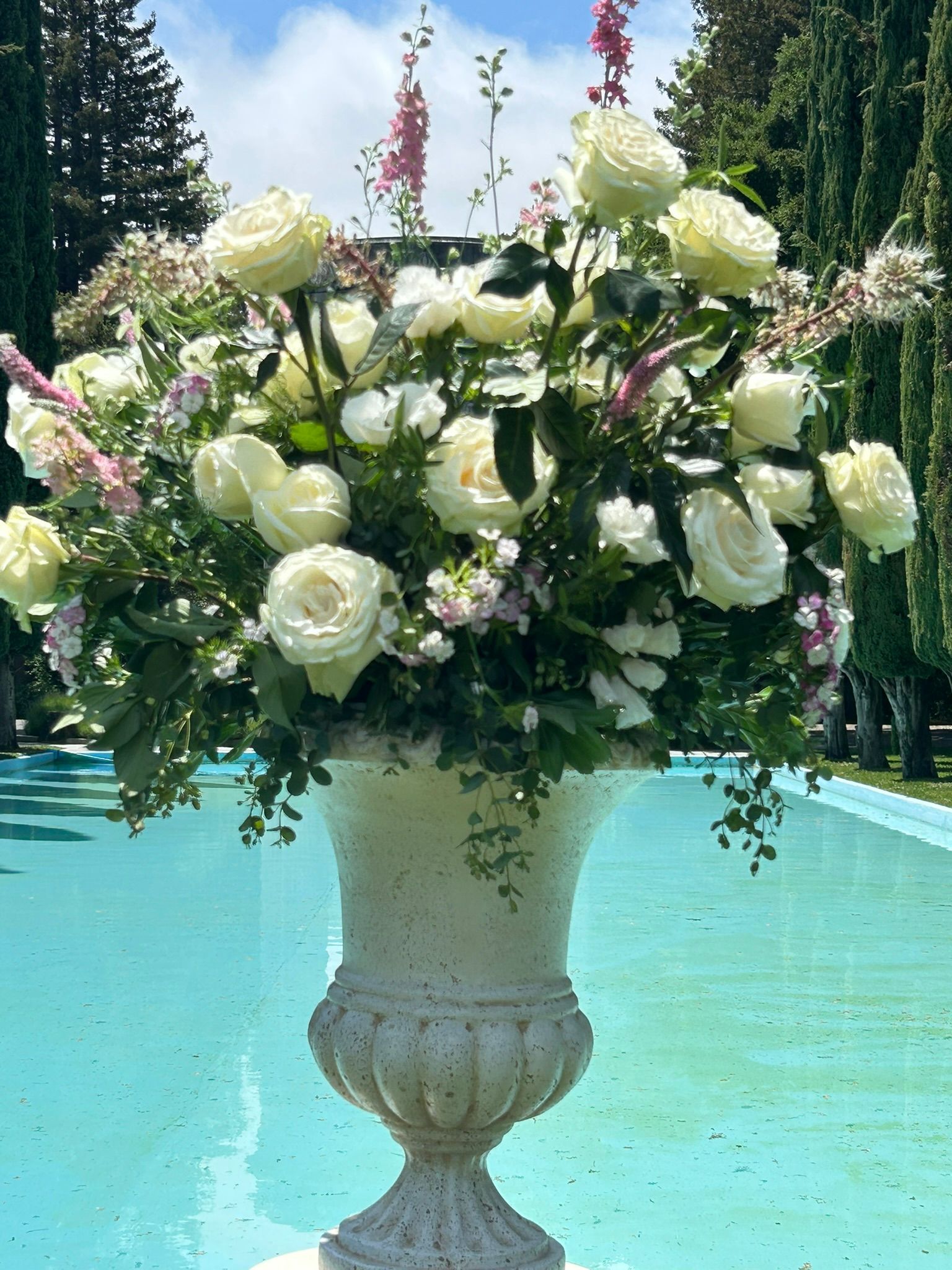 White roses and other flowers in a decorative urn by a turquoise pool.