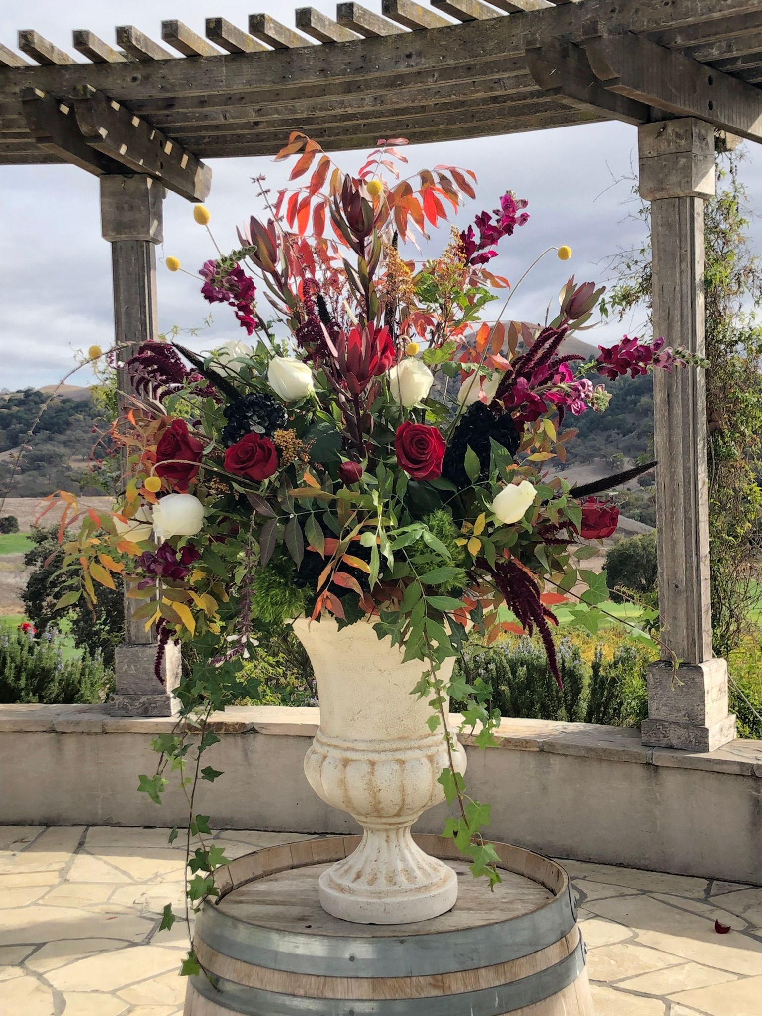 A large urn filled with burgundy and white flowers is sitting on top of a wooden barrel.