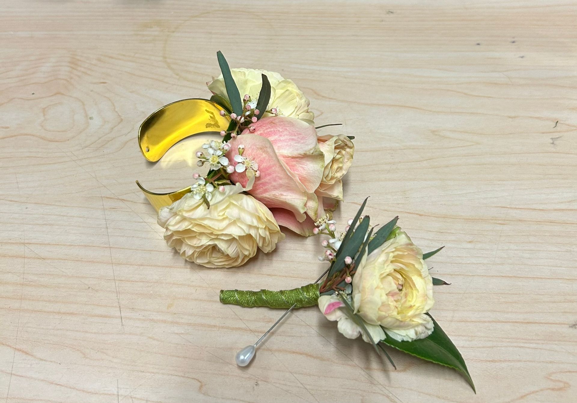 Two dried flower corsages on a wooden surface. One has a yellow ribbon, pink and cream roses. The other has a cream rose.