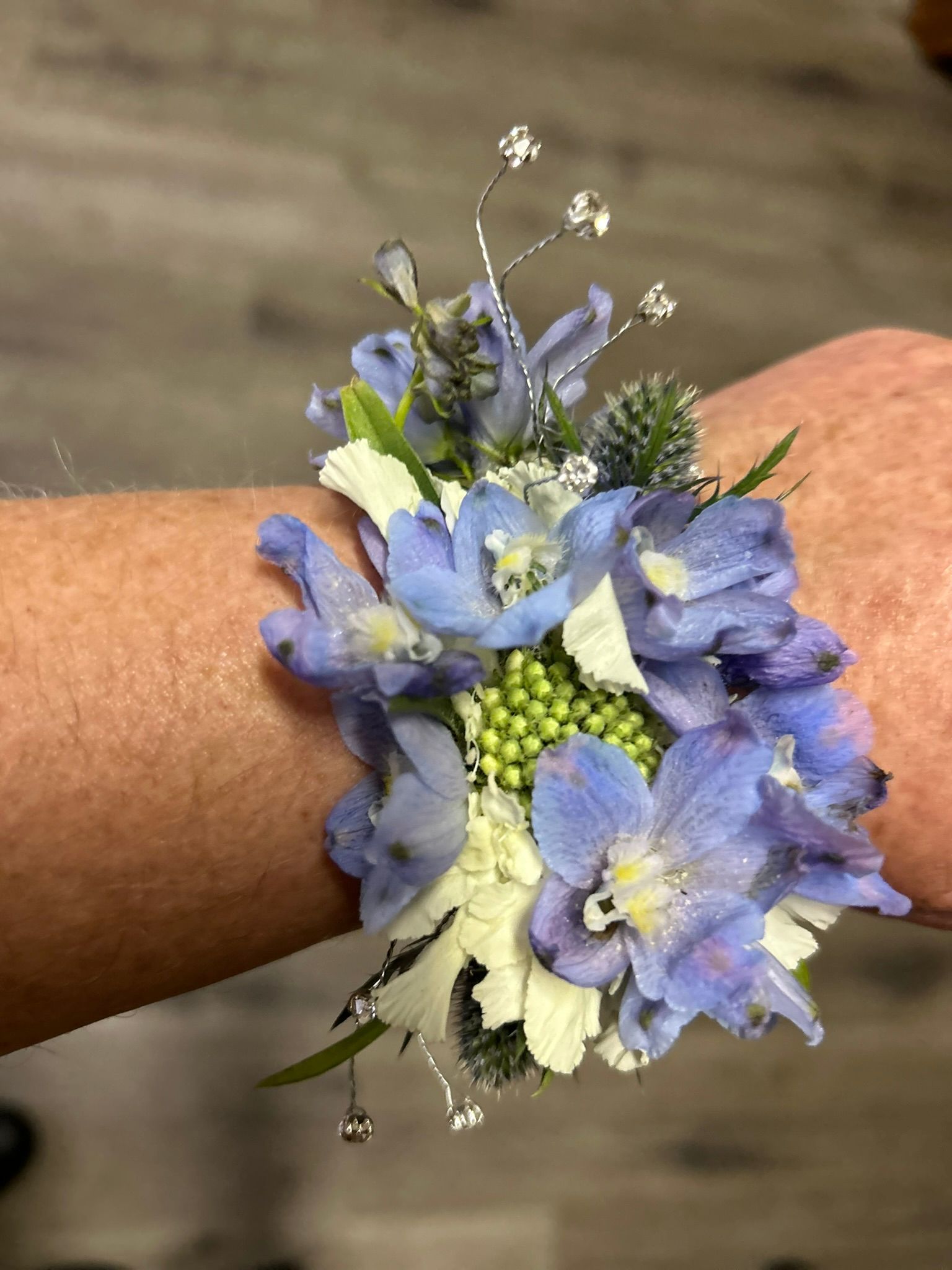 Wrist corsage with light blue and white flowers, greenery, and silver accents.