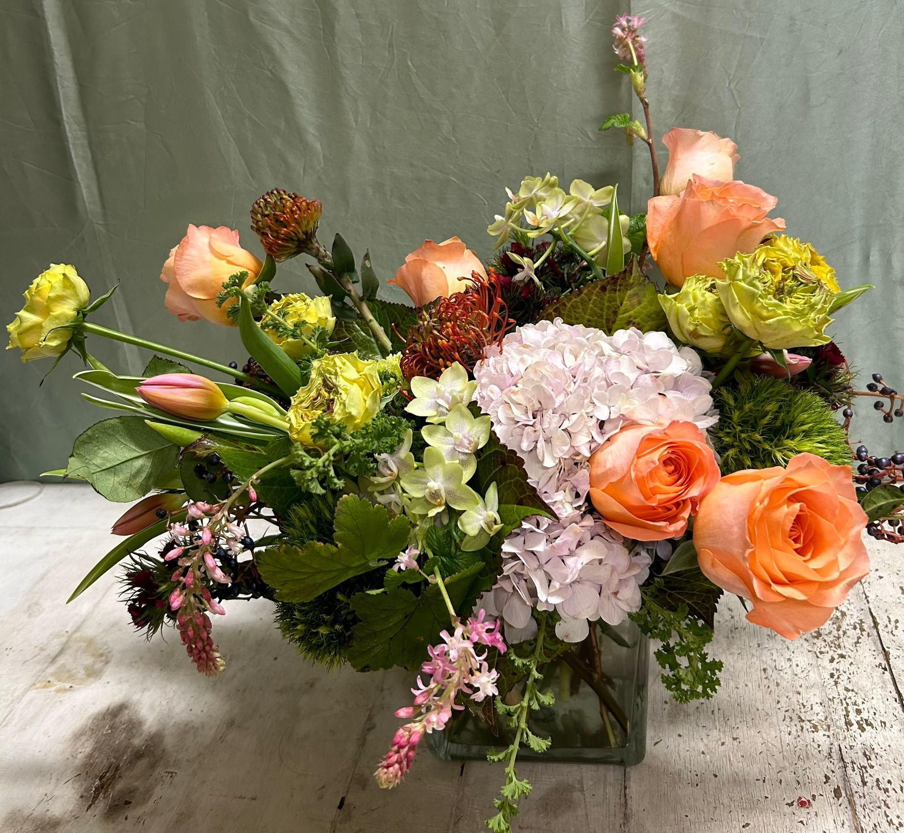 Floral arrangement in a square glass vase, featuring peach roses, hydrangeas, and various greenery.