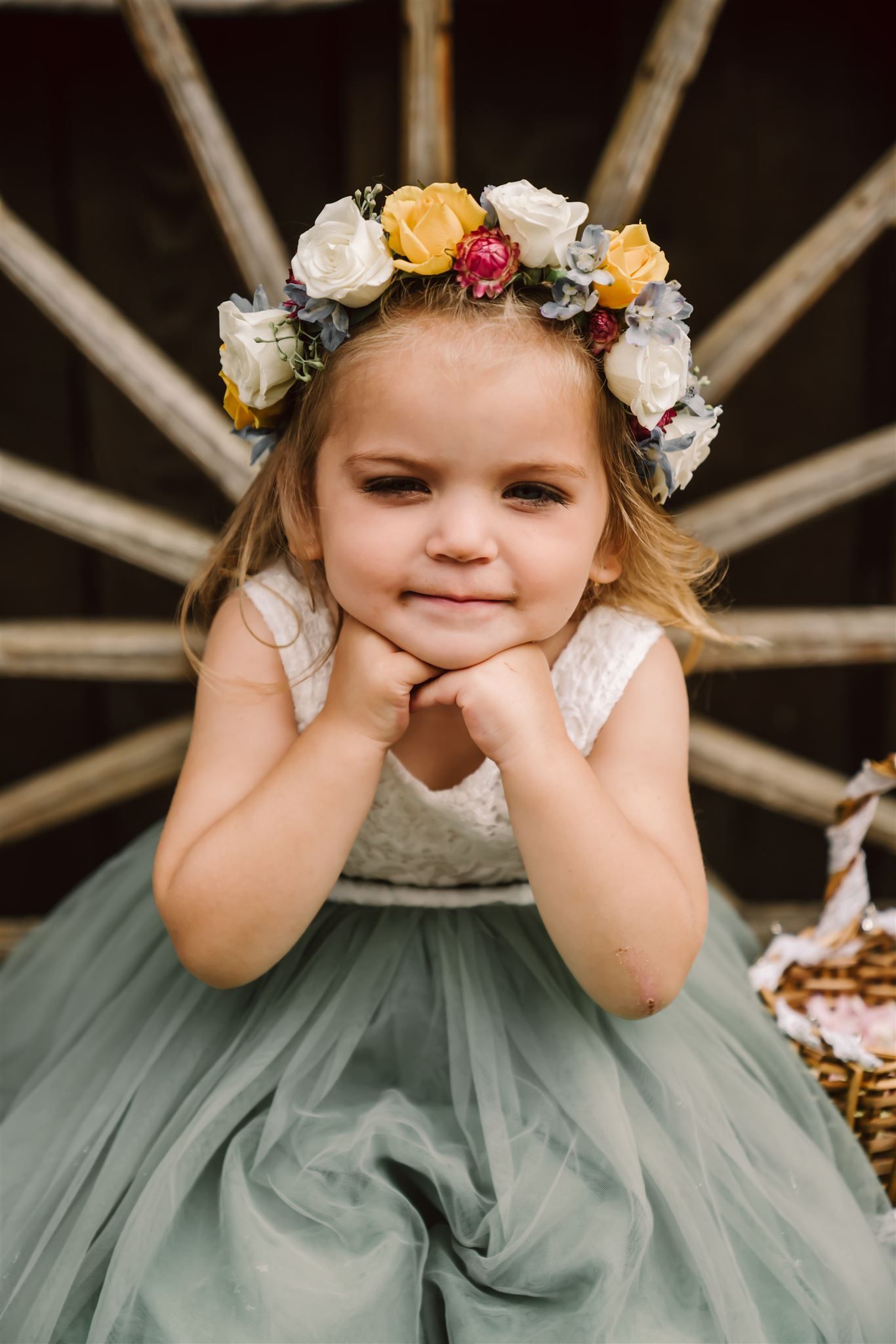 A little girl wearing a flower crown is sitting in front of a wagon wheel.