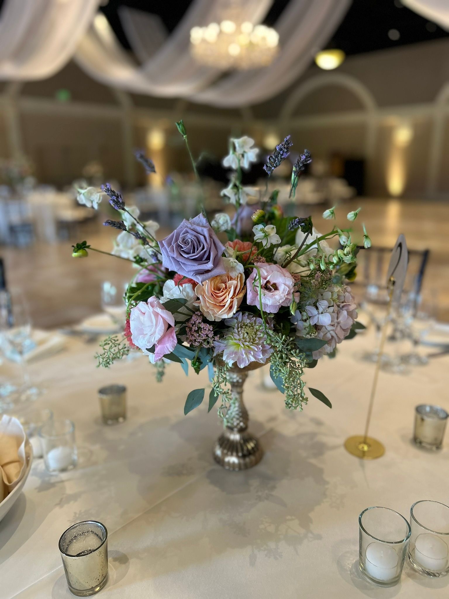 Centerpiece with purple, pink, and peach flowers in a golden vase on a table set for a wedding.