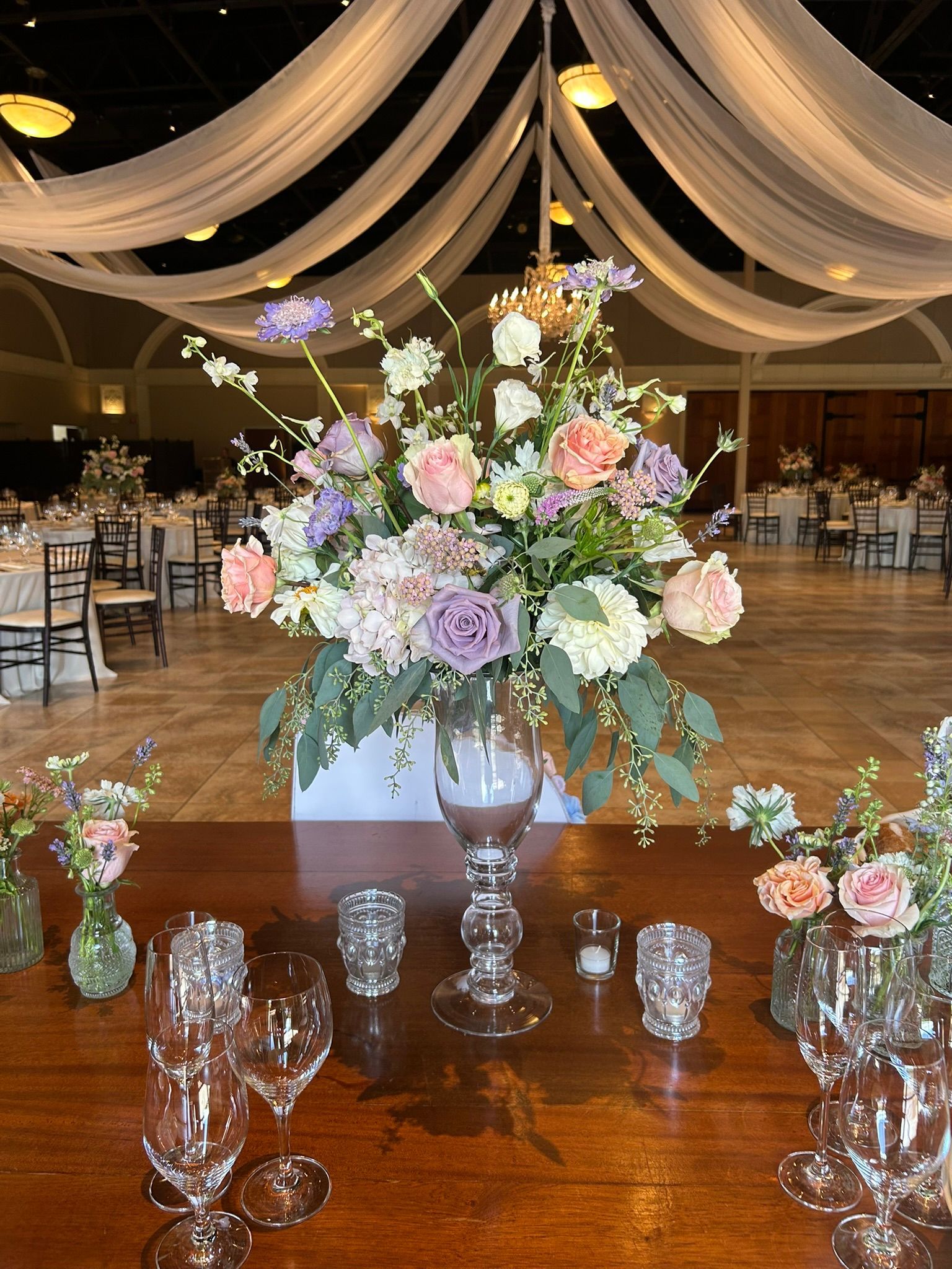 Wedding reception table with floral centerpiece, candles, and glassware. Dim lighting, white draped ceiling.