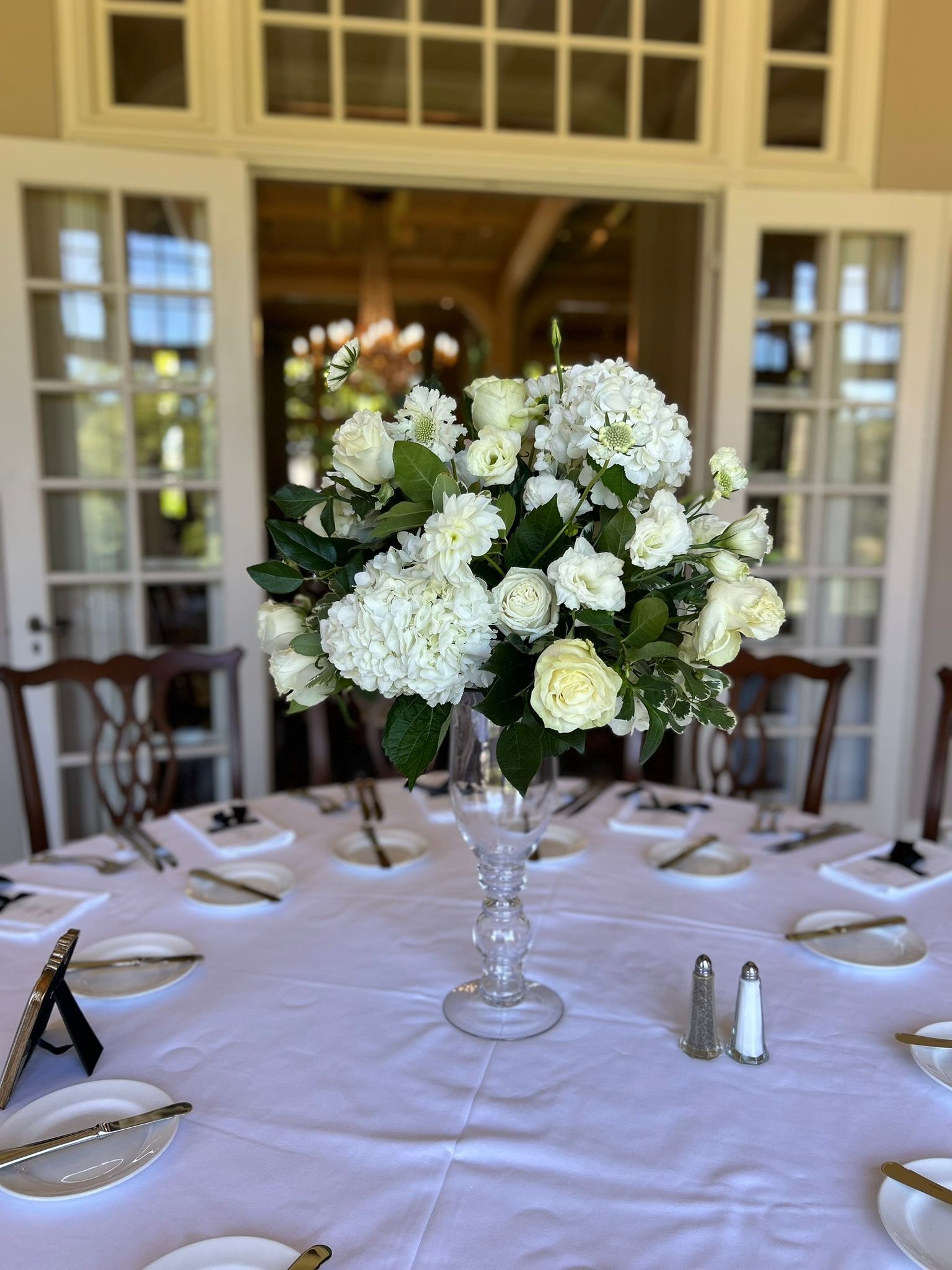 White floral centerpiece on a round table set for dining, with an open French door background.