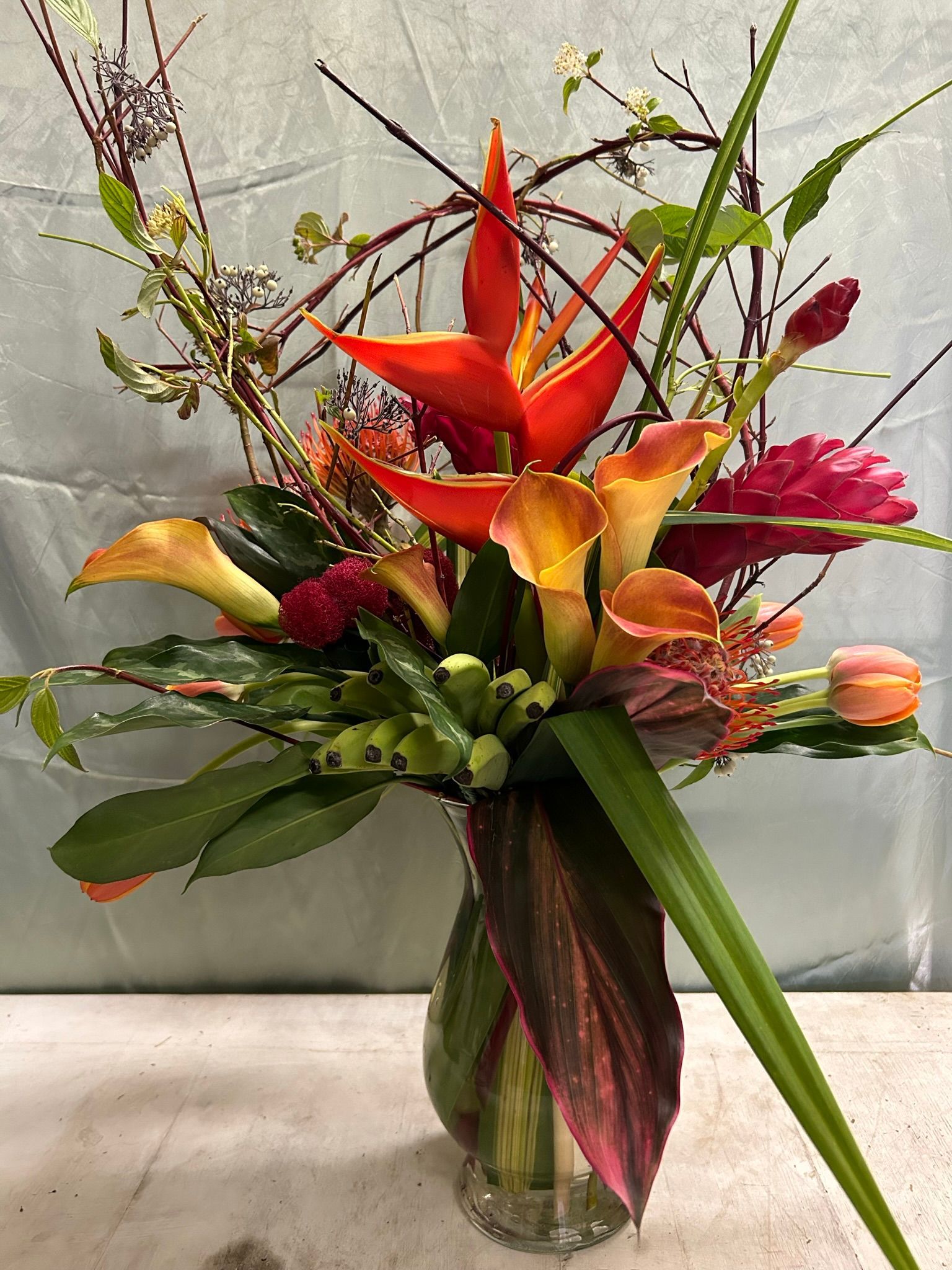 A vase filled with tropical flowers that include Heliconia, calla lilies, ginger and branches