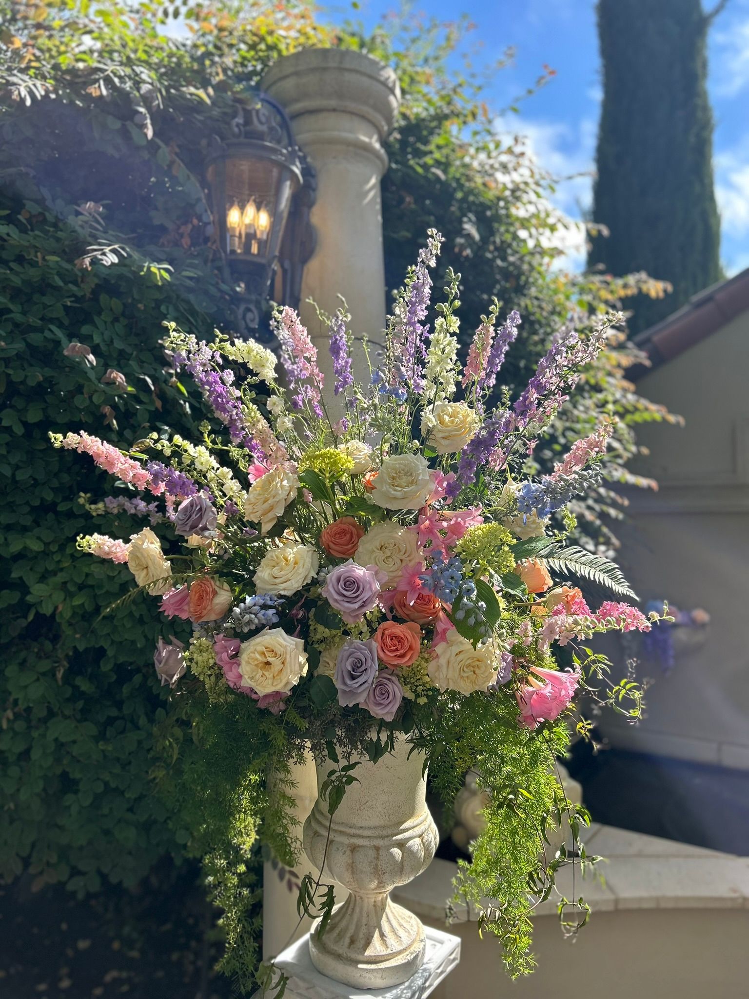 Urn overflowing with pastel flowers, set against a green hedge and stone pillar in a sunny outdoor space.