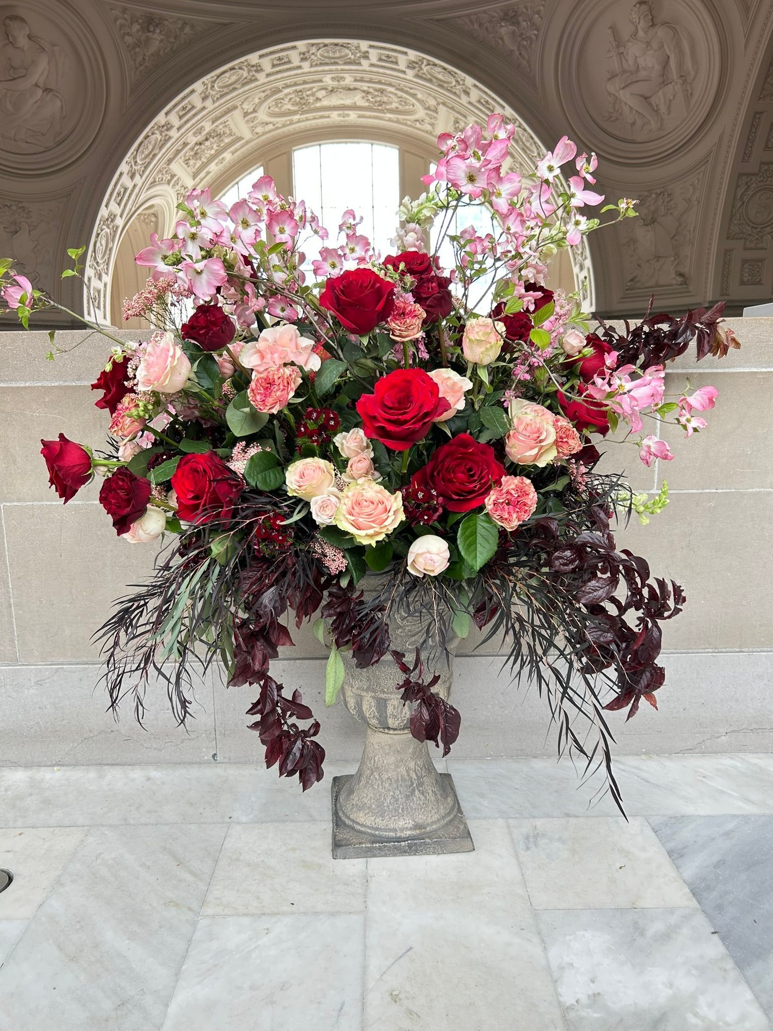 Large floral arrangement of red and pink flowers in a stone urn, set against an architectural backdrop.