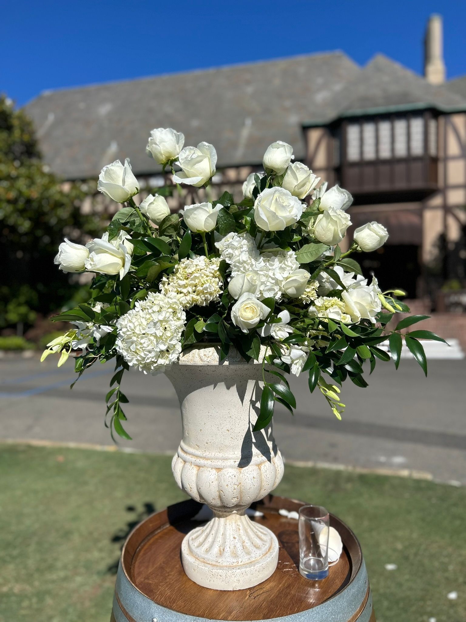White floral arrangement in urn on a barrel; Tudor-style building in background.