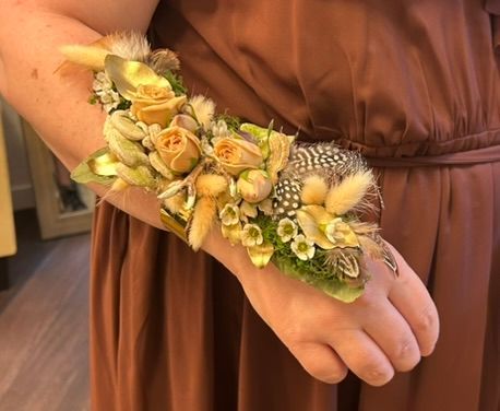 Wrist corsage of yellow roses, feathers, and greenery on a woman's wrist, wearing a brown dress.