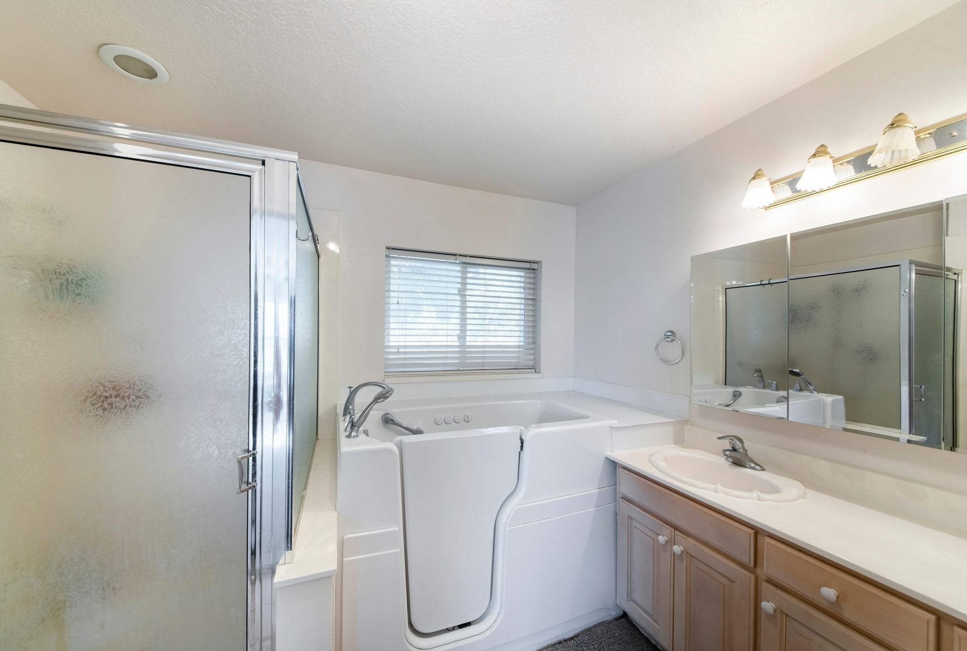 Bathroom with a walk-in tub and a frosted glass shower, white walls, and light wood vanity.