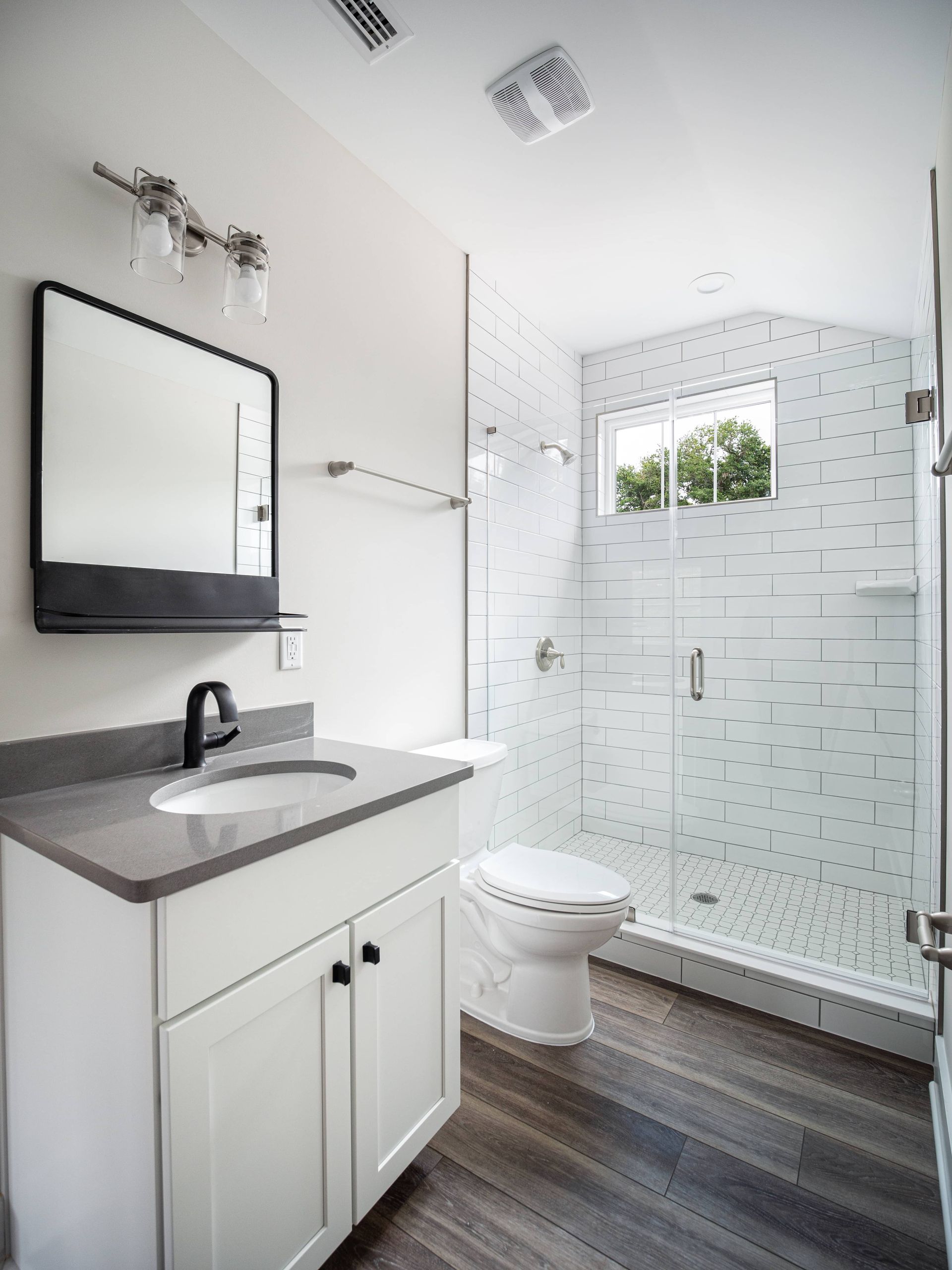 Modern white bathroom with gray countertop, dark wood floor, glass shower, and toilet.