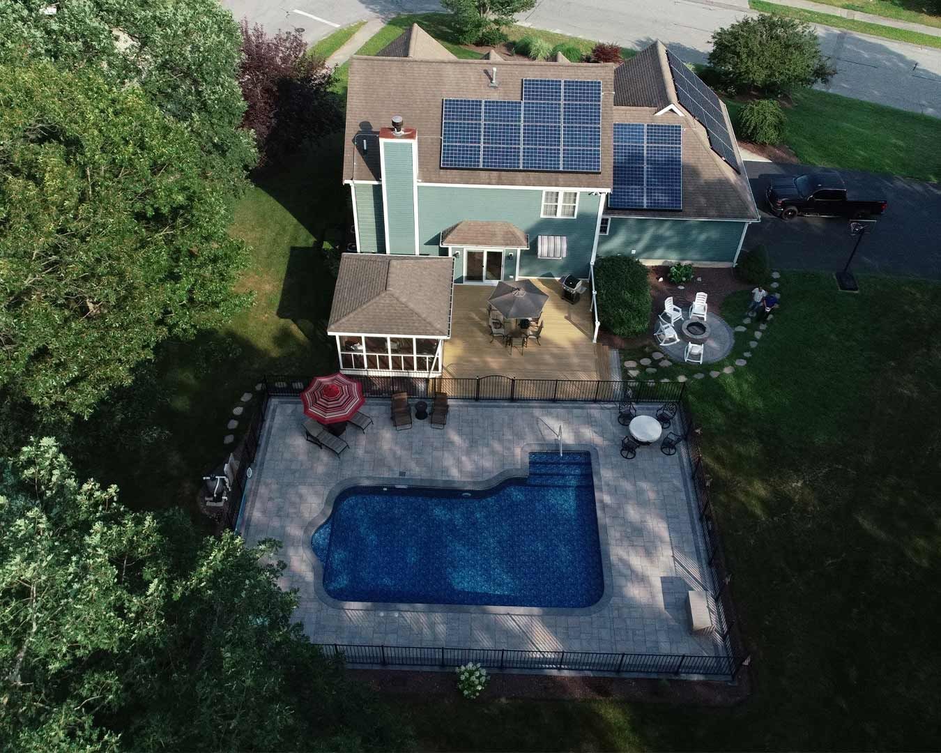 Aerial view of a teal house with solar panels, a backyard patio, and a swimming pool surrounded by a fence and trees.