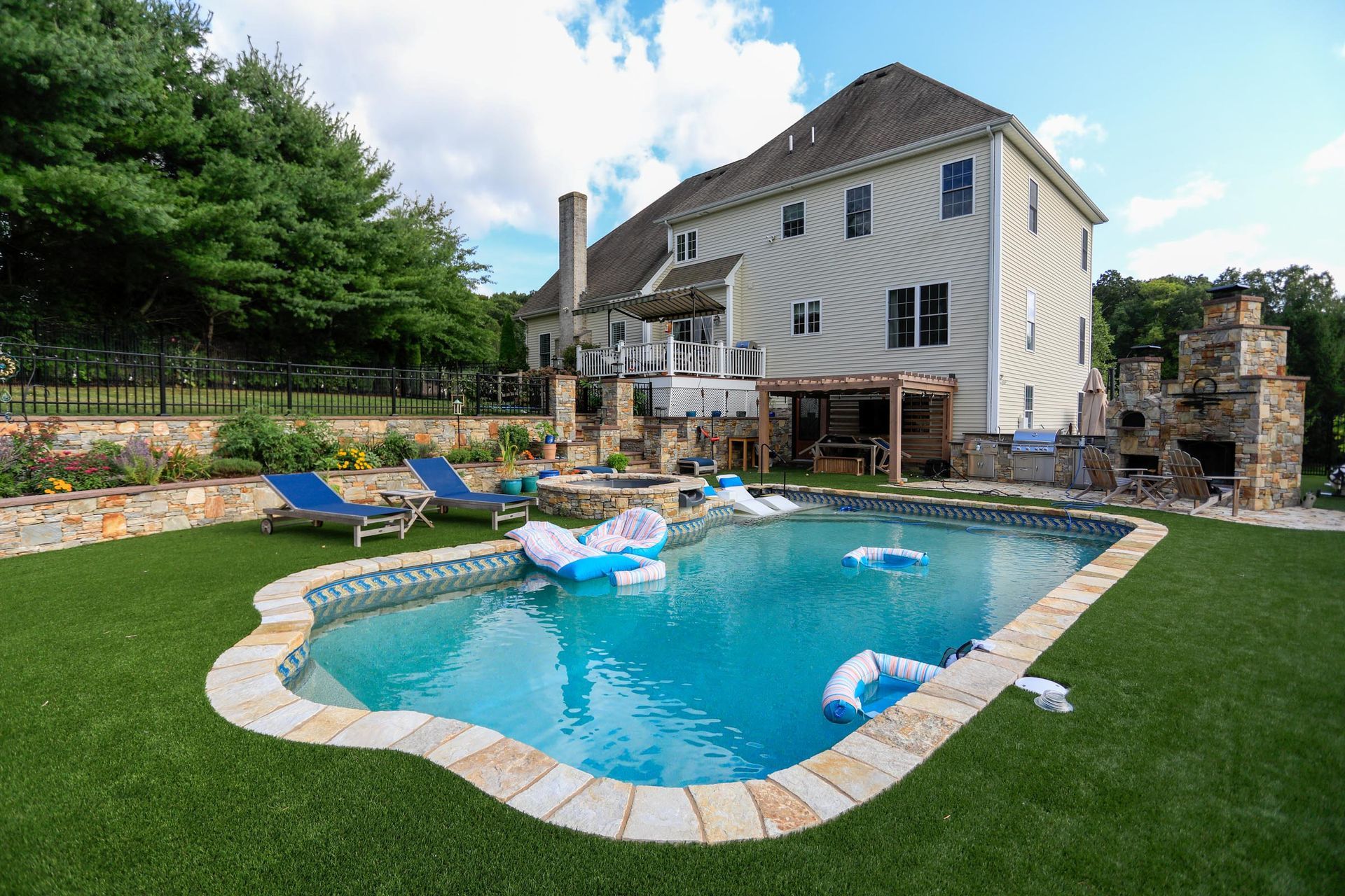 A large backyard with a swimming pool, lounge chairs, a stone outdoor fireplace, and a beige house under a blue sky.