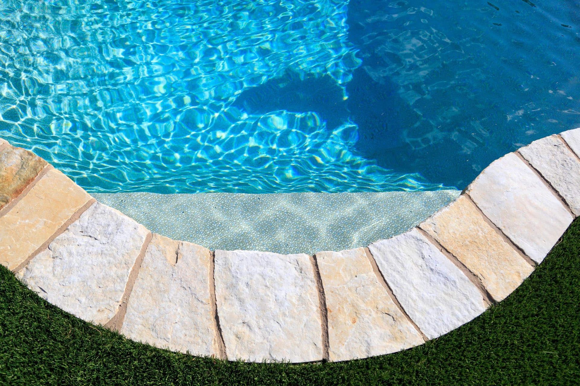 A curved stone pool deck bordering clear blue water and a lush green lawn.