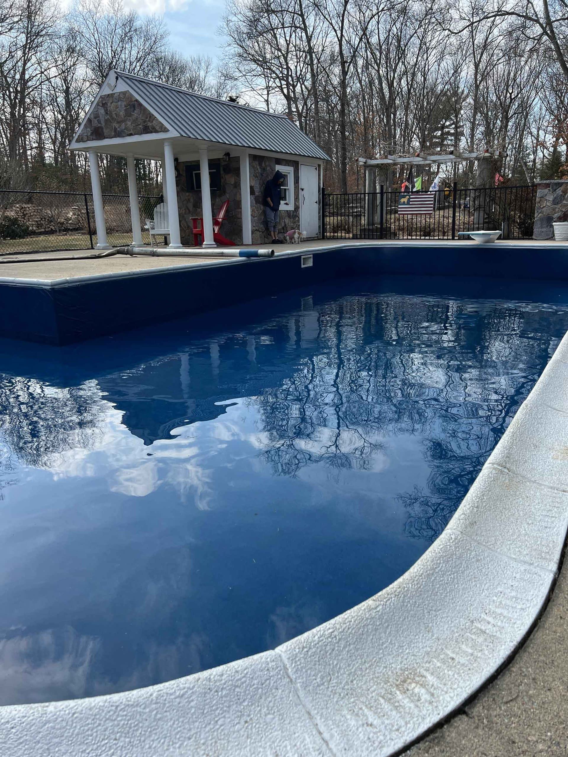 A deep blue swimming pool with a textured white stone edge, set before a small gazebo in a wooded backyard.