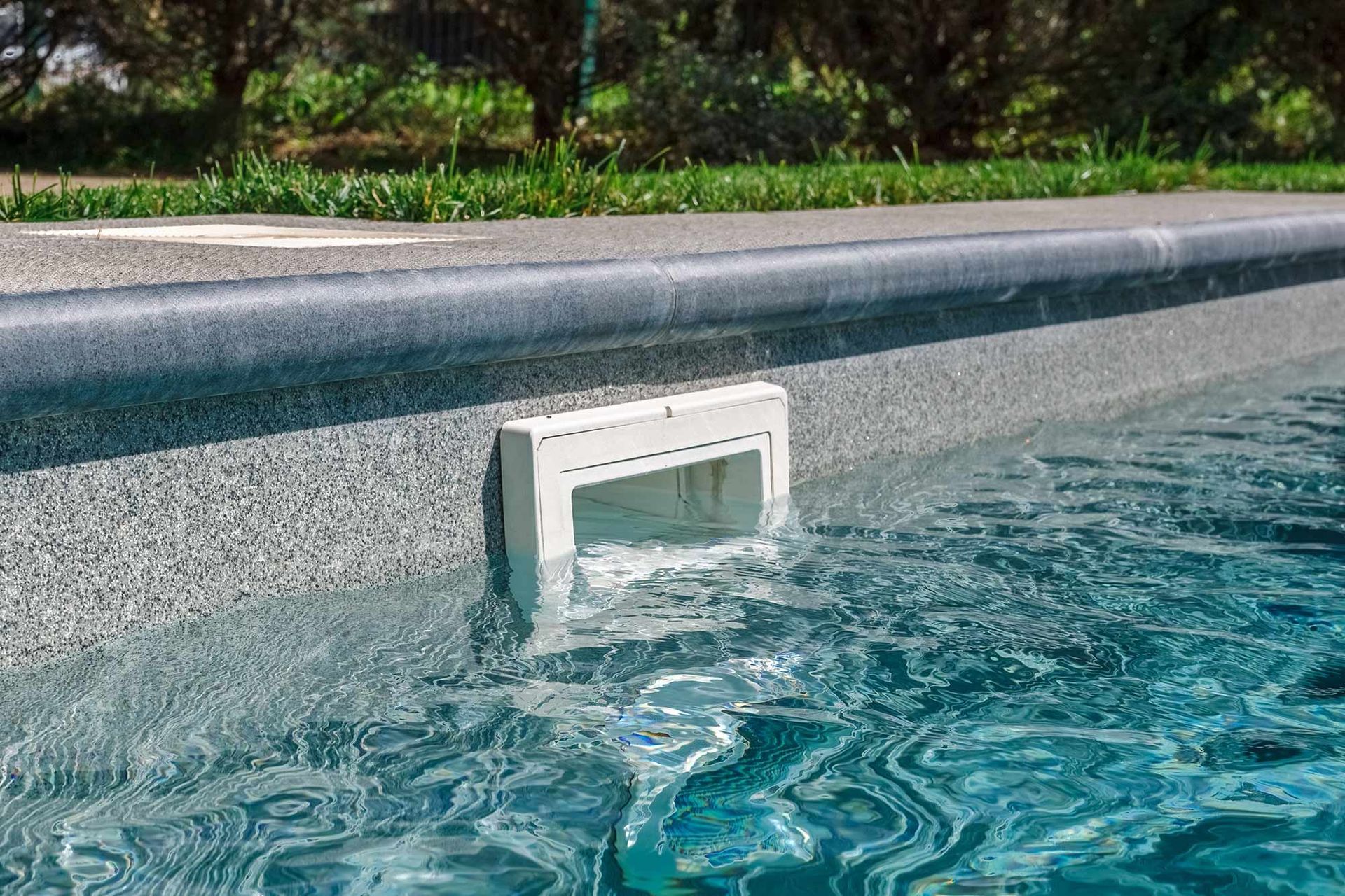 A white pool skimmer box installed into the grey, textured wall of a swimming pool at the water line.