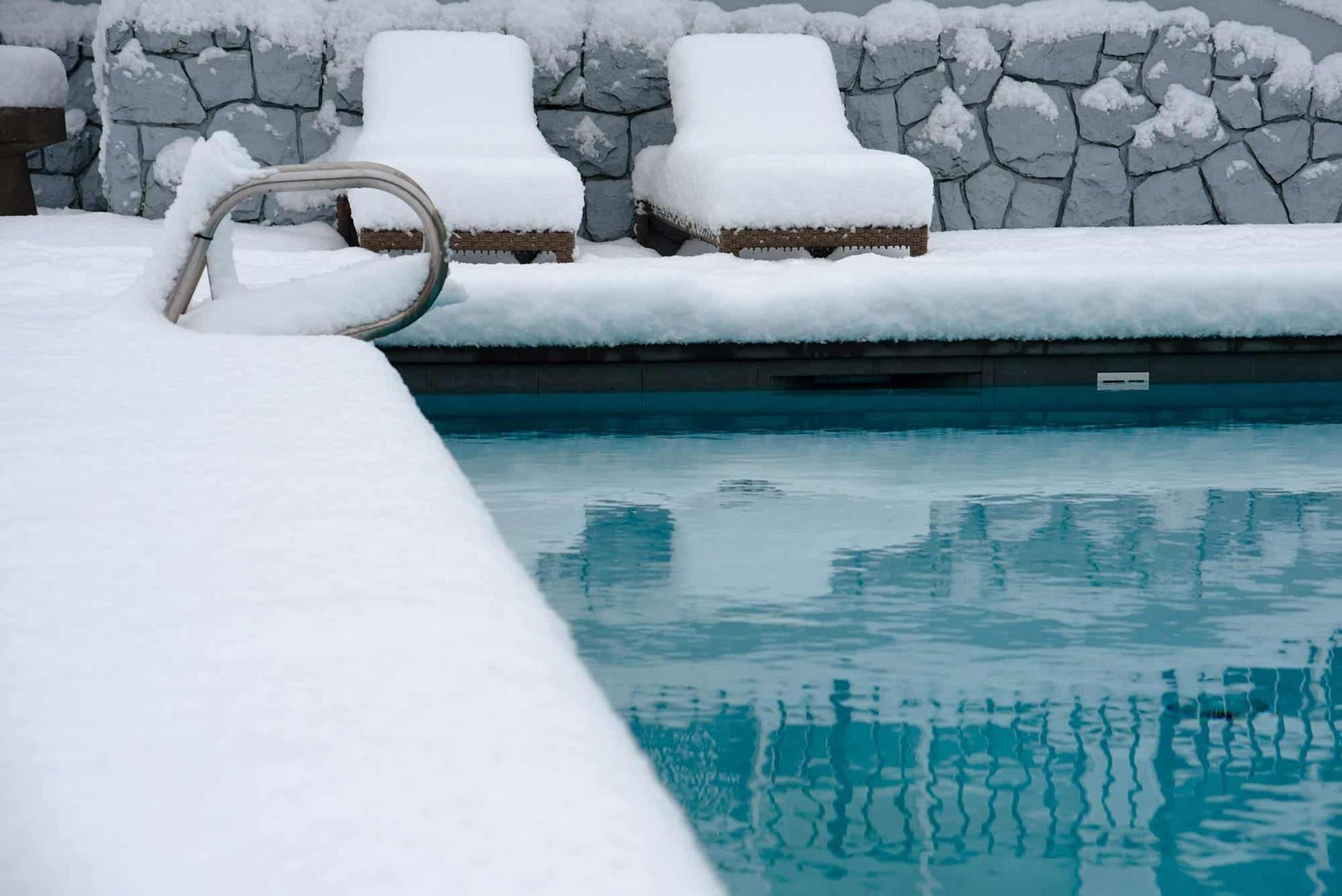 Two snow-covered lounge chairs sit by a clear blue swimming pool surrounded by a stone wall covered in fresh snow.