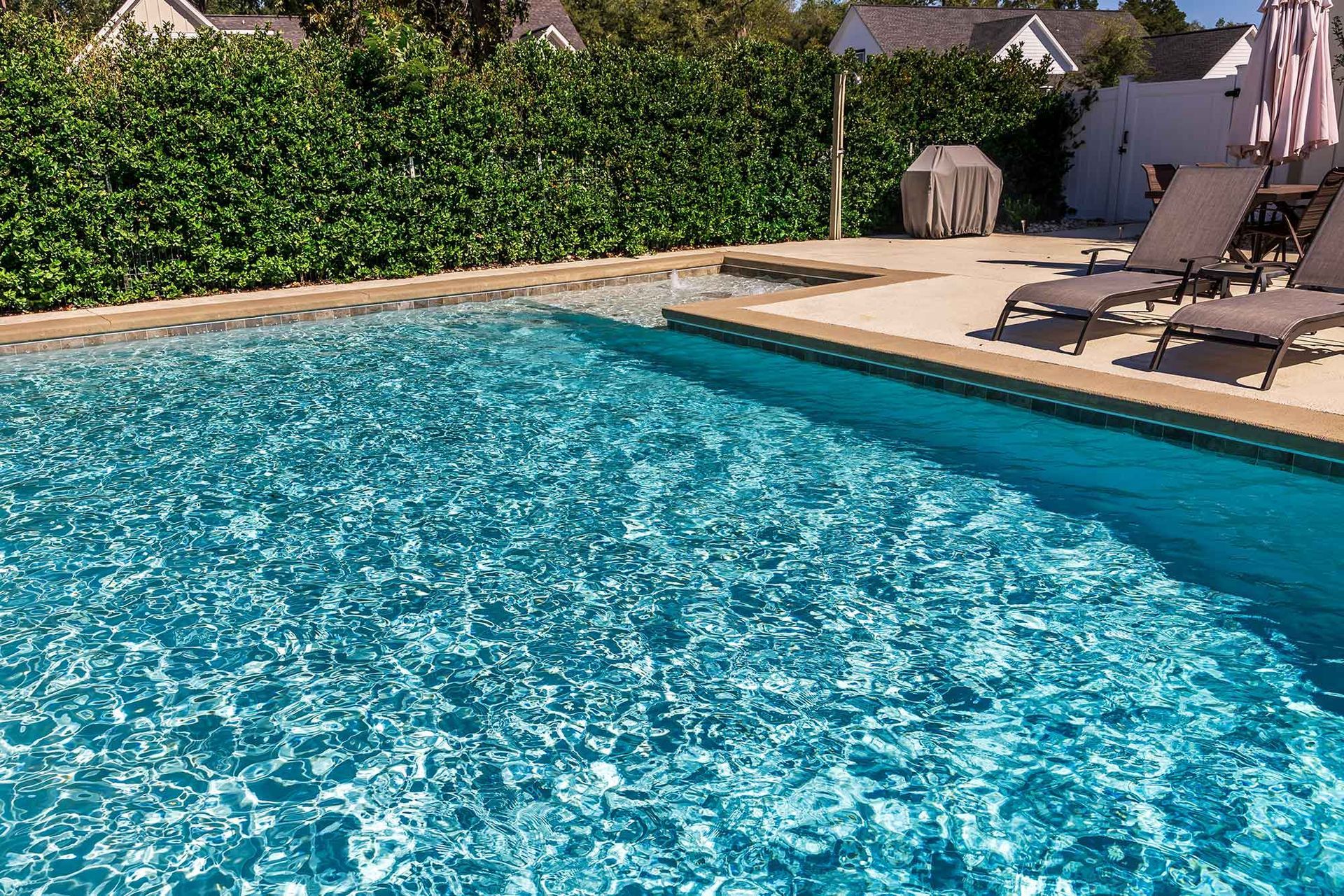 A bright blue swimming pool with a stone deck, lounge chairs, and a lush green hedge in a sunny outdoor setting.
