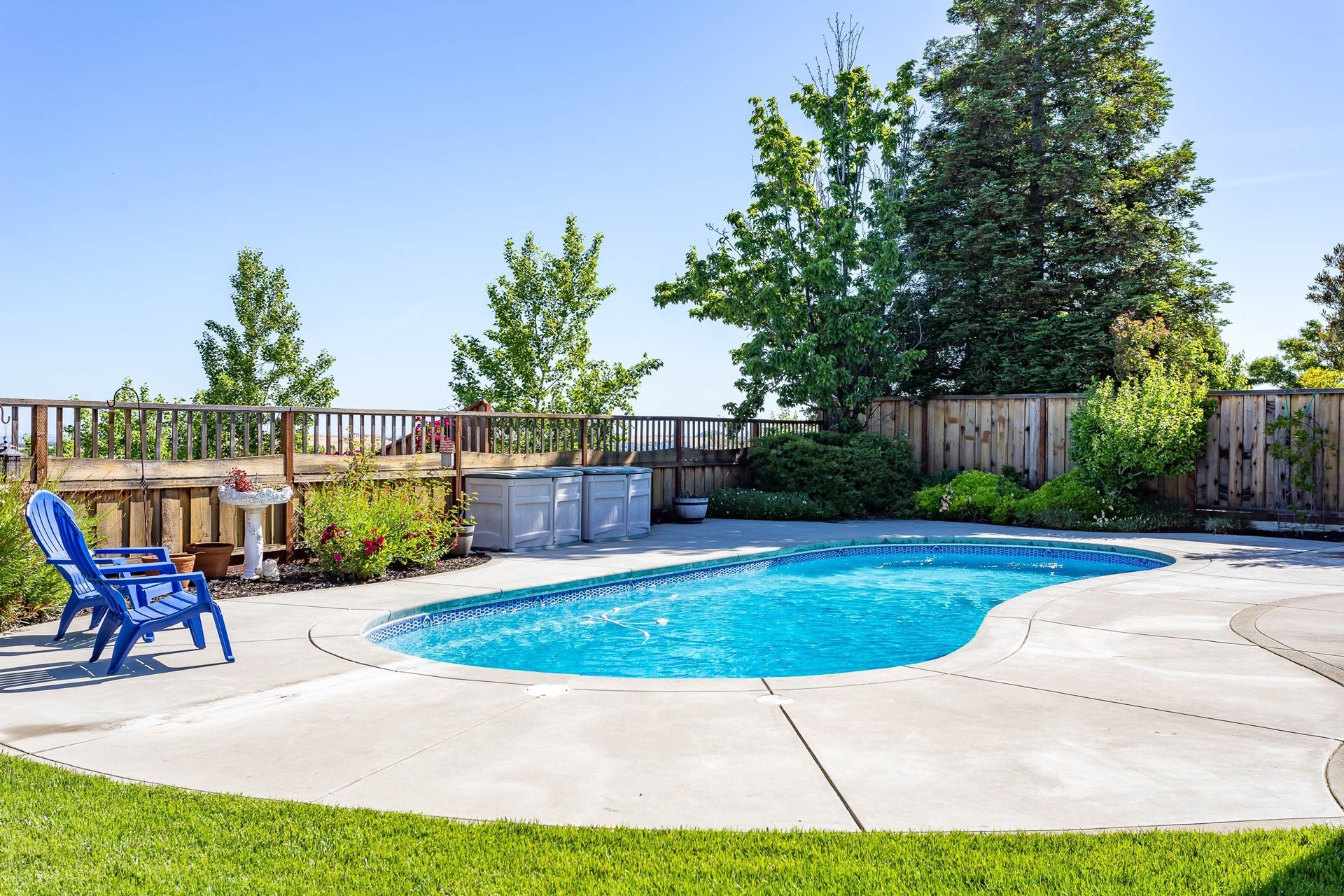 A bright blue backyard swimming pool with a concrete deck, two blue chairs, and lush green trees under a clear blue sky.