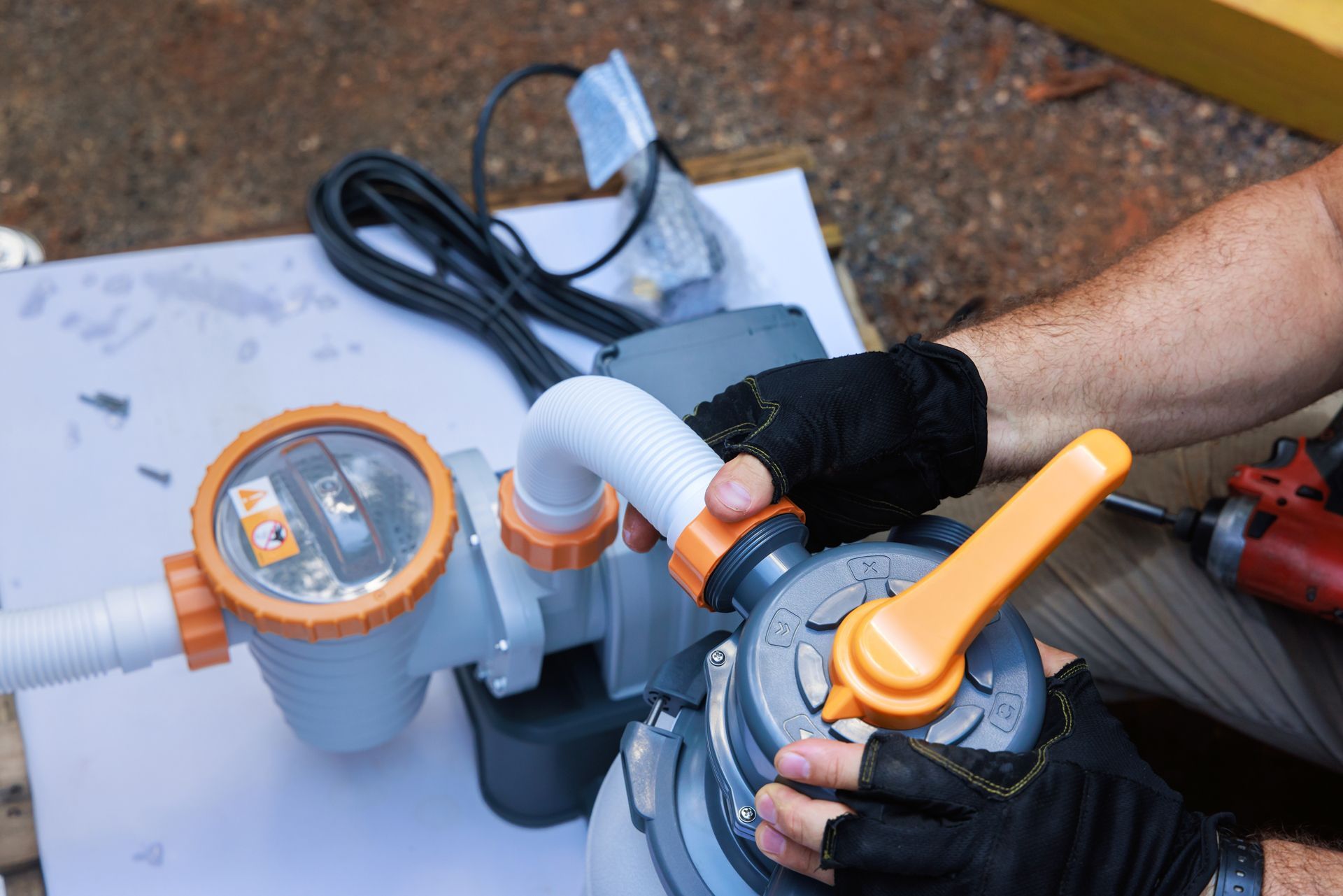 A person wearing black gloves installs a white hose onto a blue and orange pool pump system outdoors.
