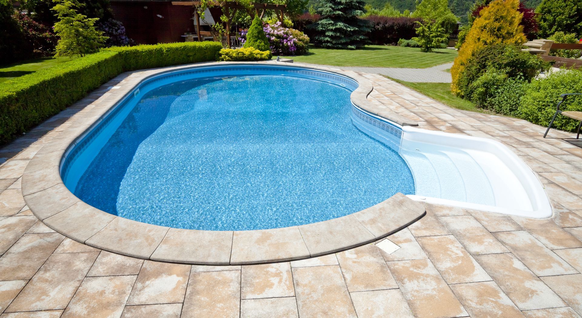 A kidney-shaped swimming pool with blue water and white entry steps, surrounded by stone pavers and a green hedge.