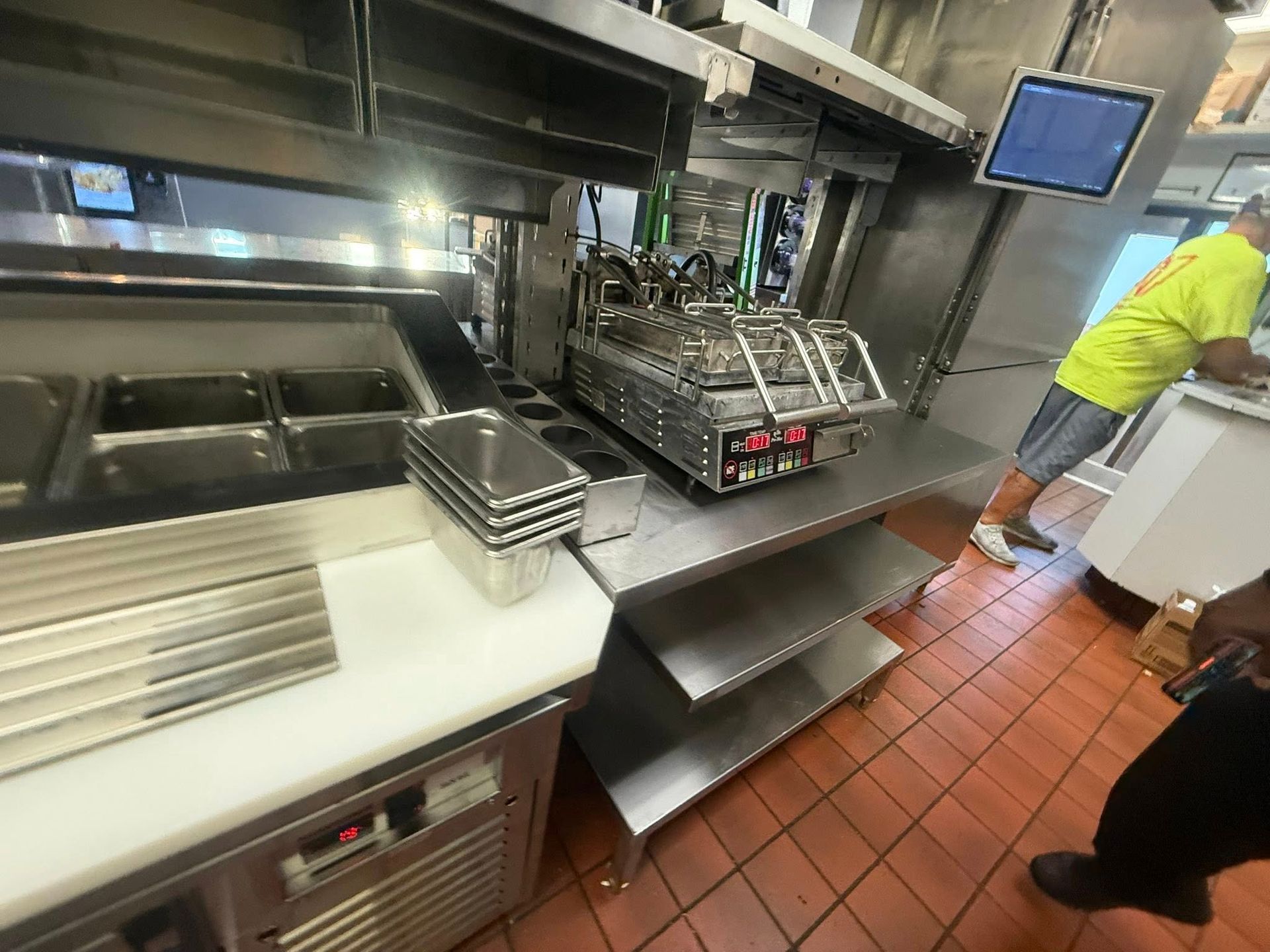 Commercial kitchen with stainless steel equipment and a worker in a yellow shirt.