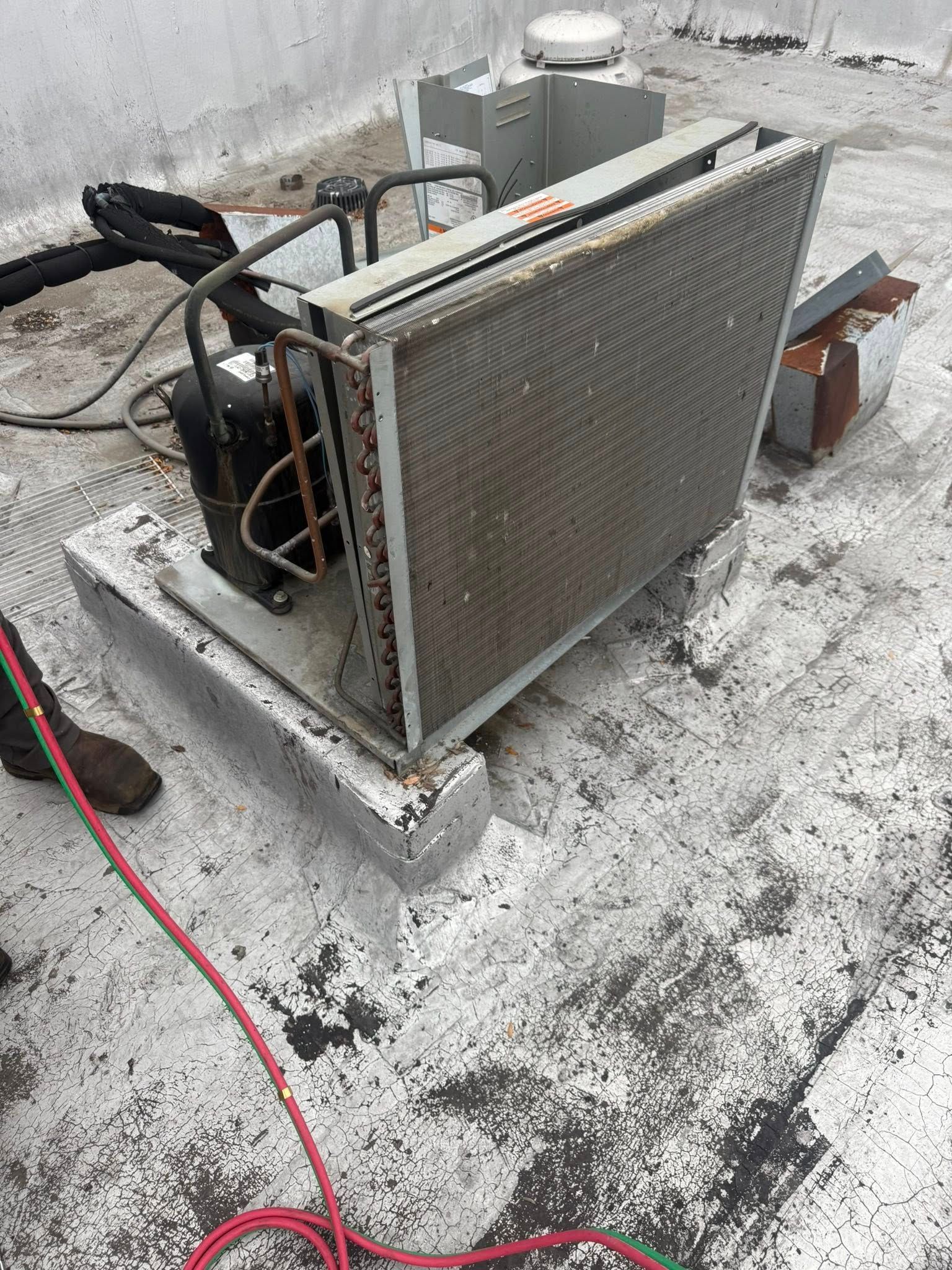 HVAC unit on a rooftop. Gray metal structure, condenser coil, black tubing, red and black cable on the ground, and white rooftop.