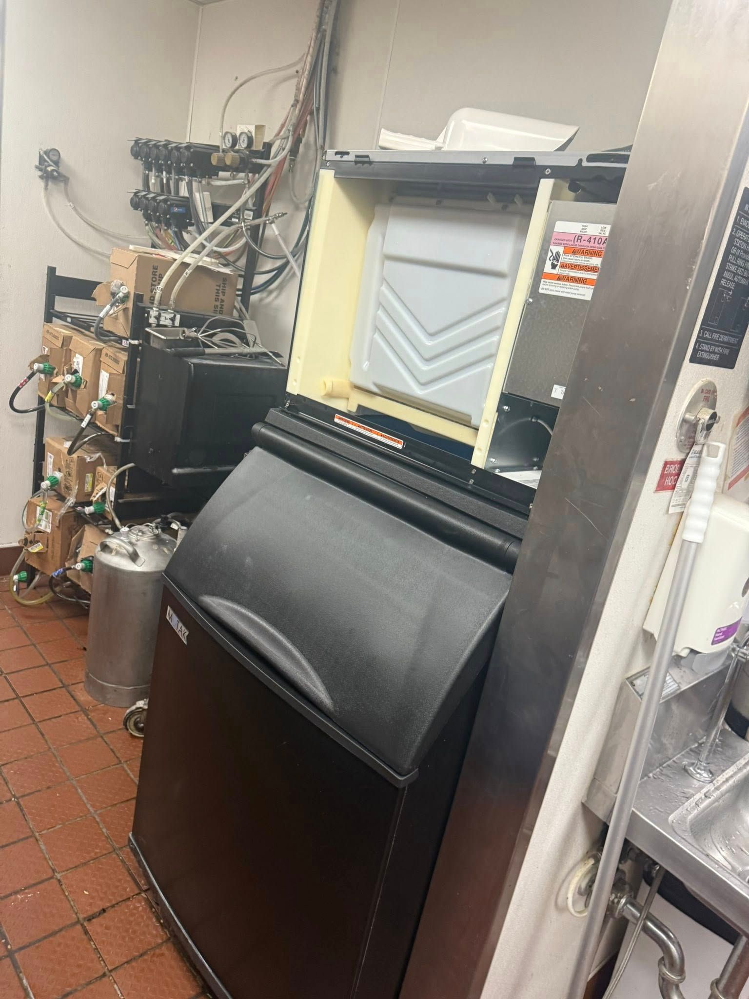 Commercial kitchen: ice machine, equipment, a metal sink, and various pipes against a wall.