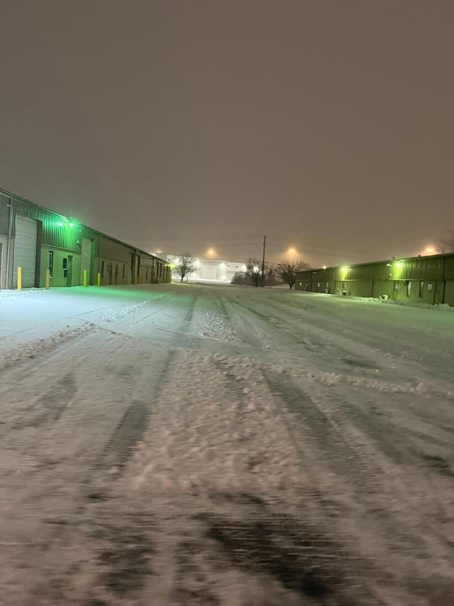 Snow-covered industrial area at night. Buildings line both sides of a snowy road, lit by green and white lights.