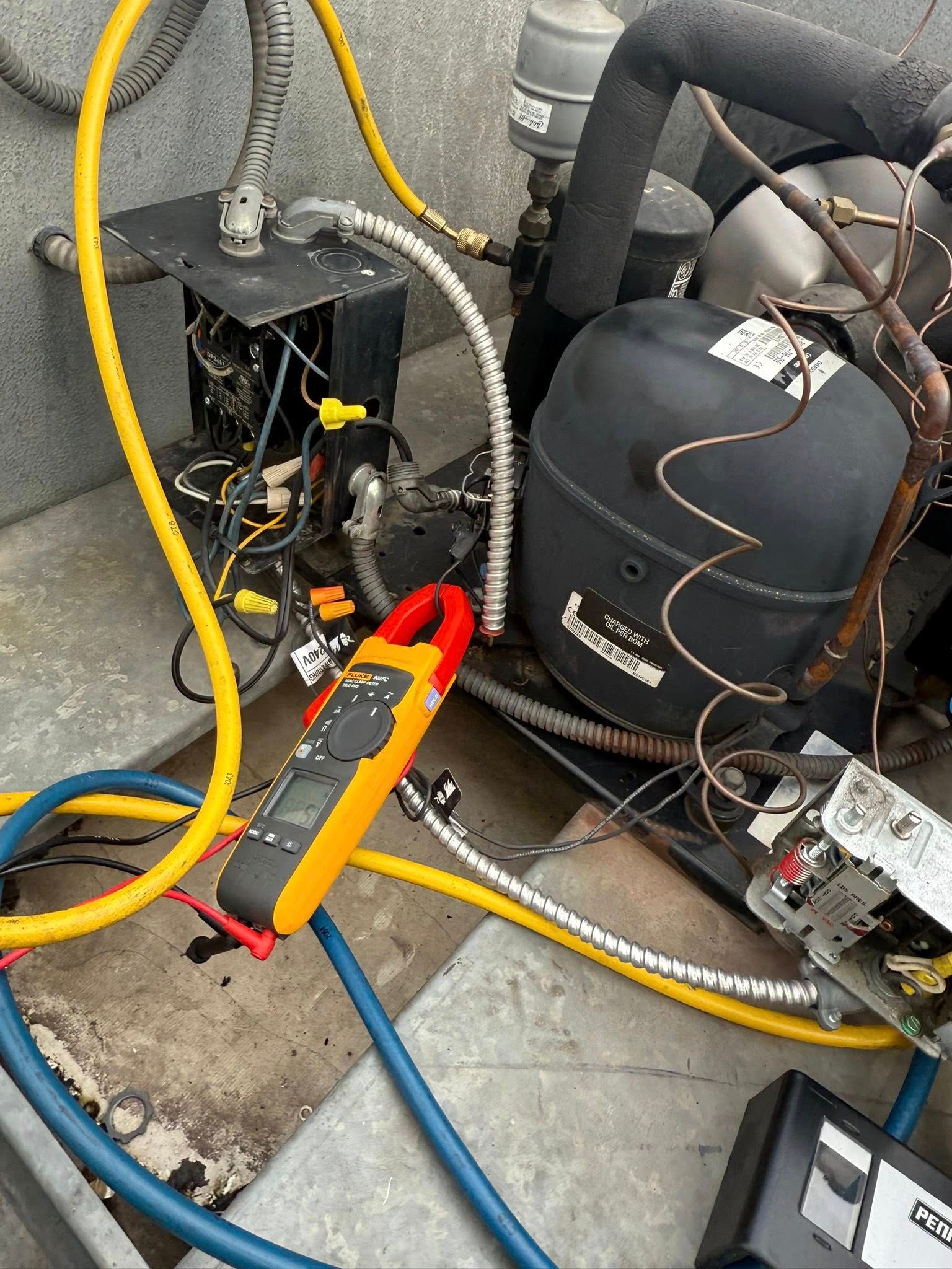 A technician uses a clamp meter on refrigeration equipment with yellow and blue tubing in a gray setting.