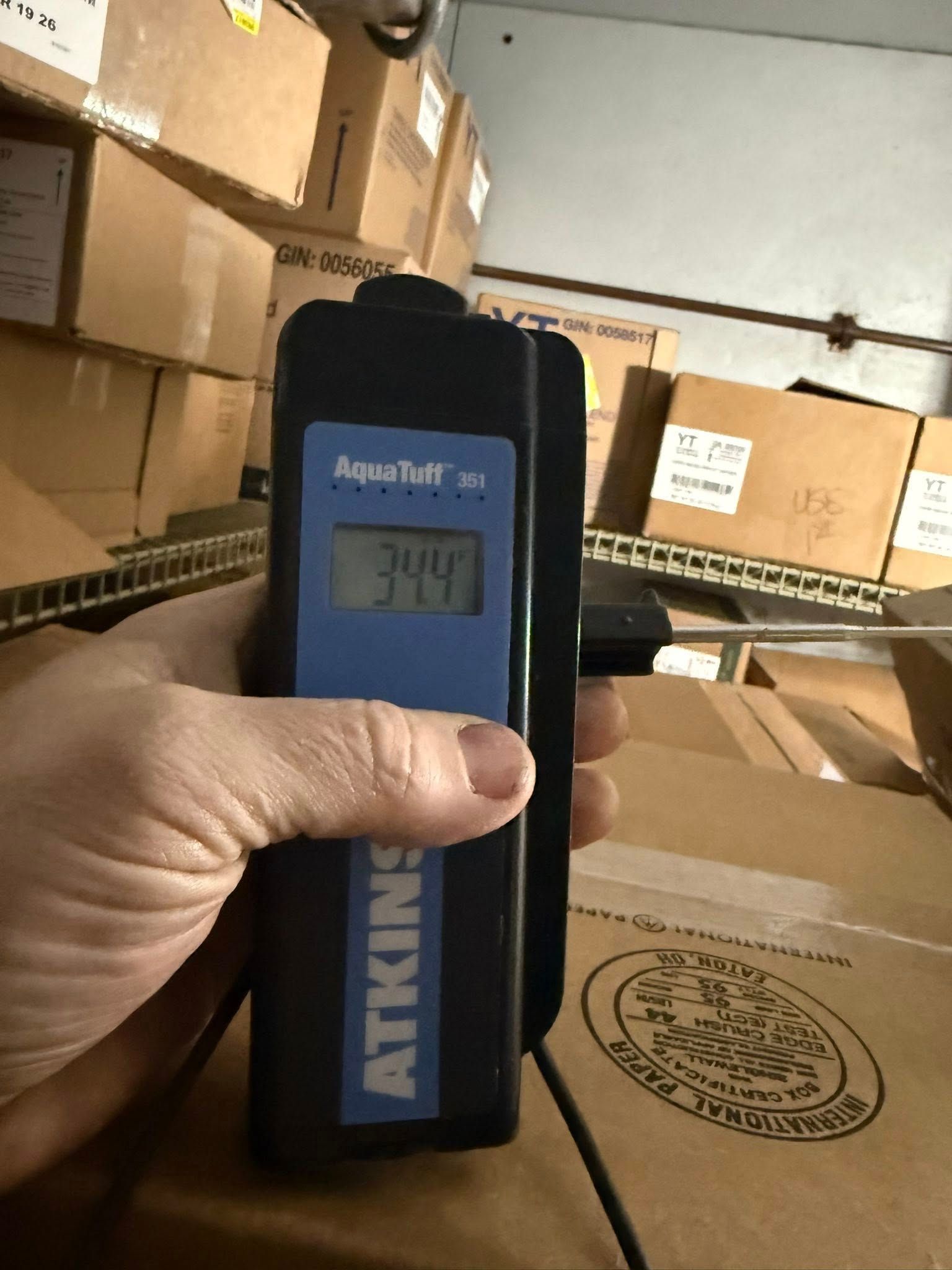 A person holds a black and blue thermometer, displaying 34.4, in a storage room with cardboard boxes.