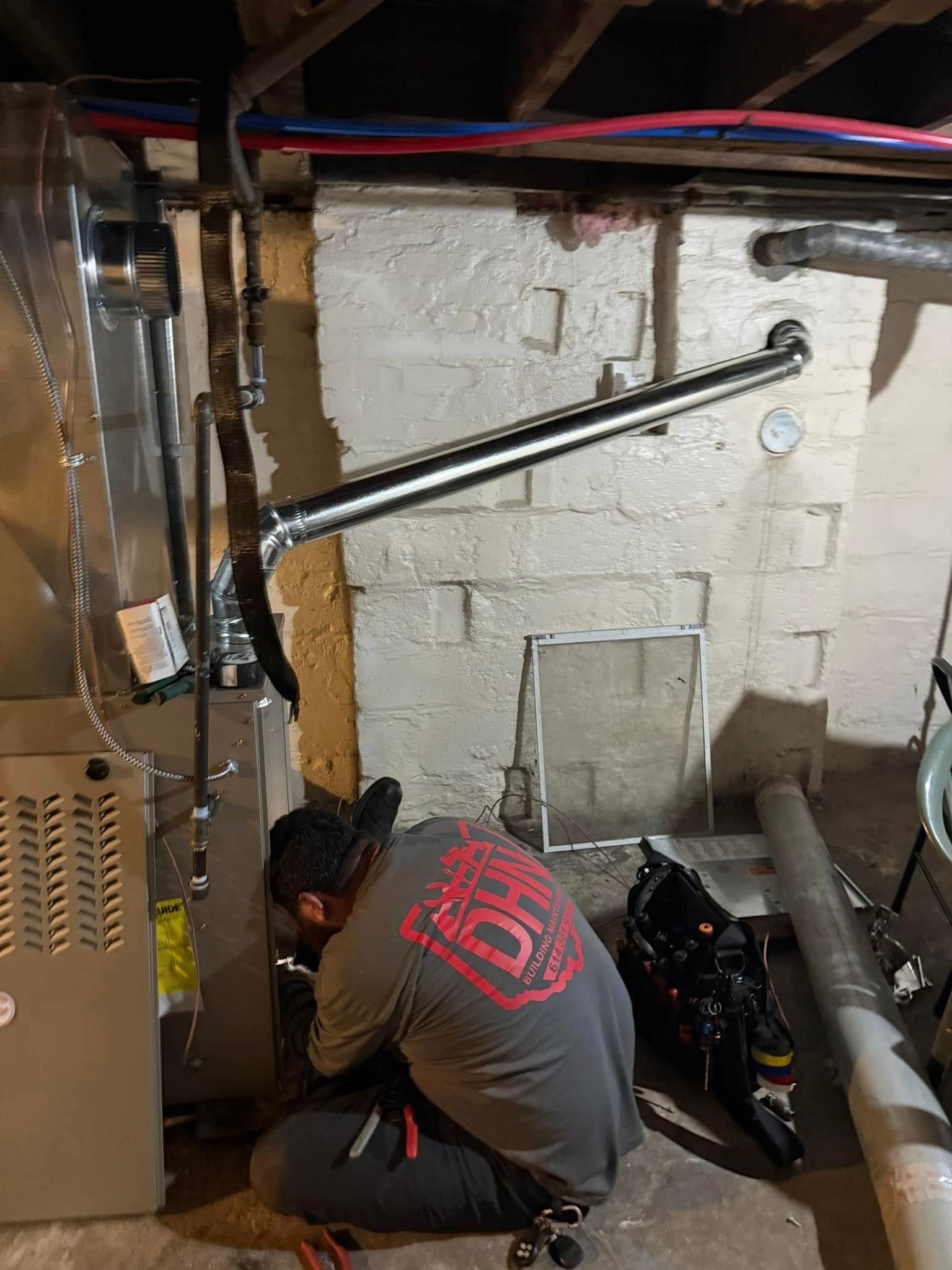 Plumber working on a furnace in a basement setting; concrete wall, pipes overhead.