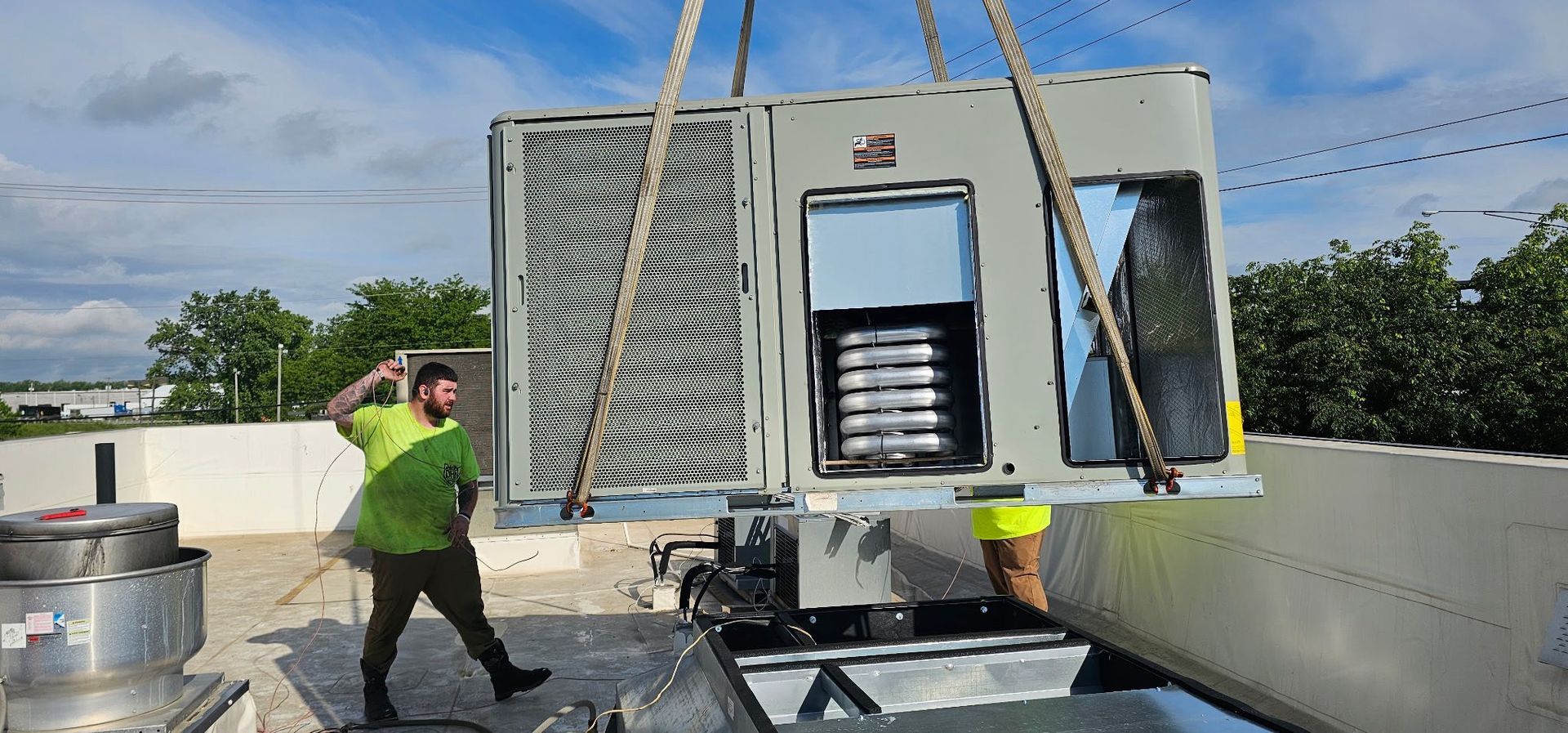 Workers installing rooftop HVAC unit. Unit suspended by ropes, men on roof directing placement.