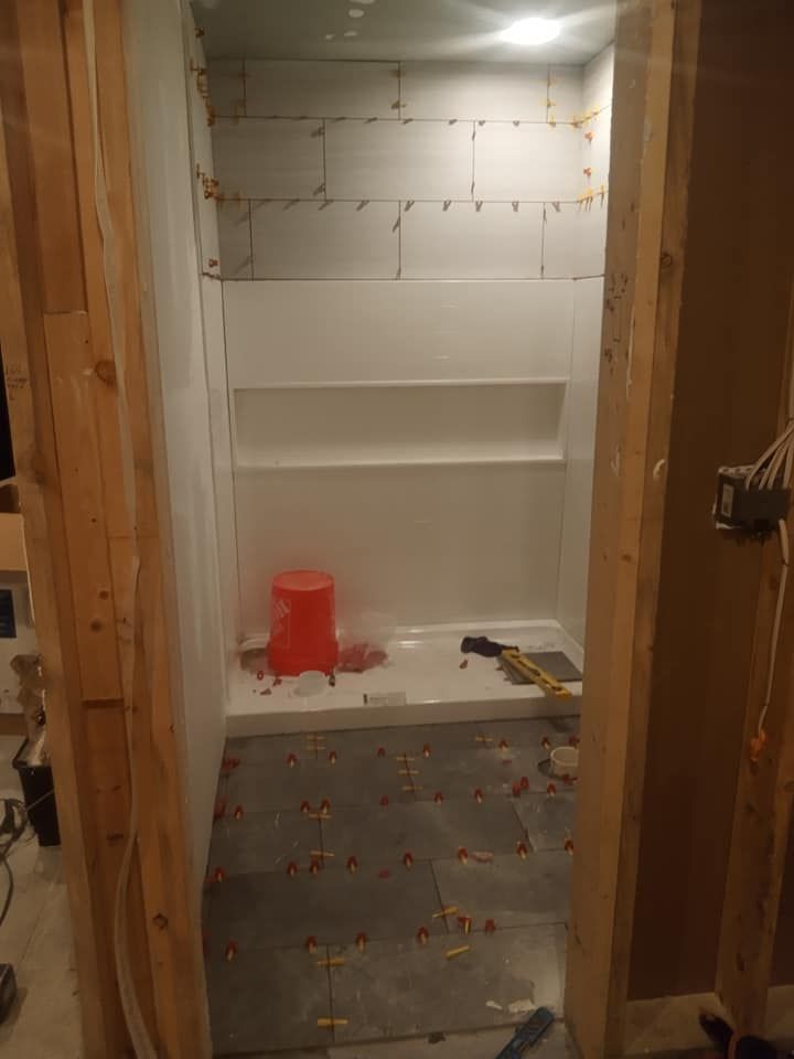 Bathroom under construction, tiled walls and floor, shower stall with a shelf, and an orange bucket.