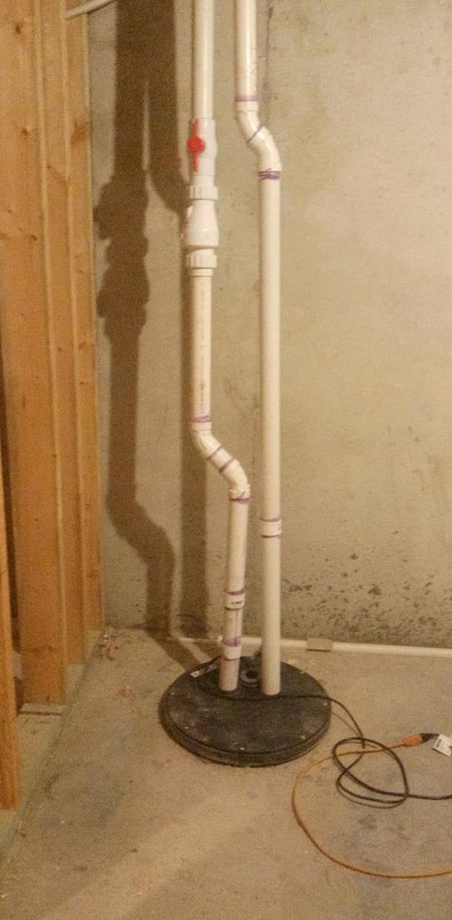 Sump pump system with pipes in a basement.  Tan pipes, gray base, concrete floor, wooden wall.