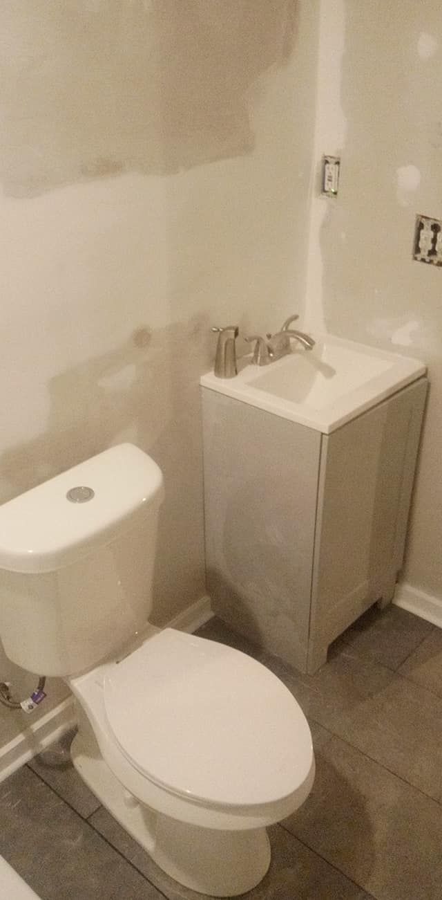 Bathroom with toilet and sink in front of white walls and gray flooring.