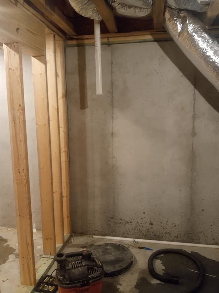 Basement corner with unfinished wooden framing, concrete wall, pipes, and sump pump.